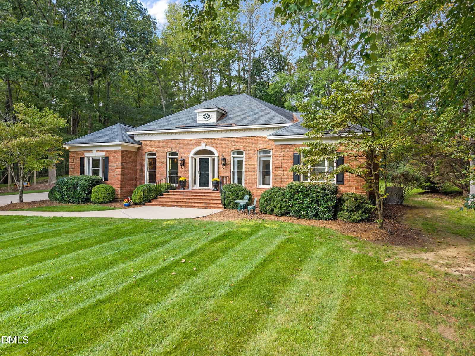 Single-story red brick house with black shutters, a gray roof, and a central entrance. The lawn is well-maintained with green grass, shrubs, and mature trees surrounding the property. Steps lead to the front door.