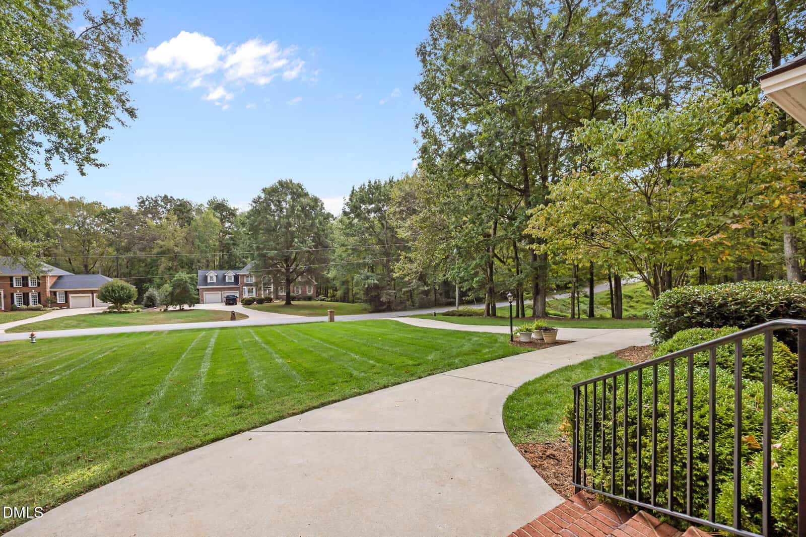 A wide concrete driveway curves through a well-manicured lawn, bordered by mature trees. Houses are visible in the background under a clear blue sky. Black railing and brick steps are in the foreground.