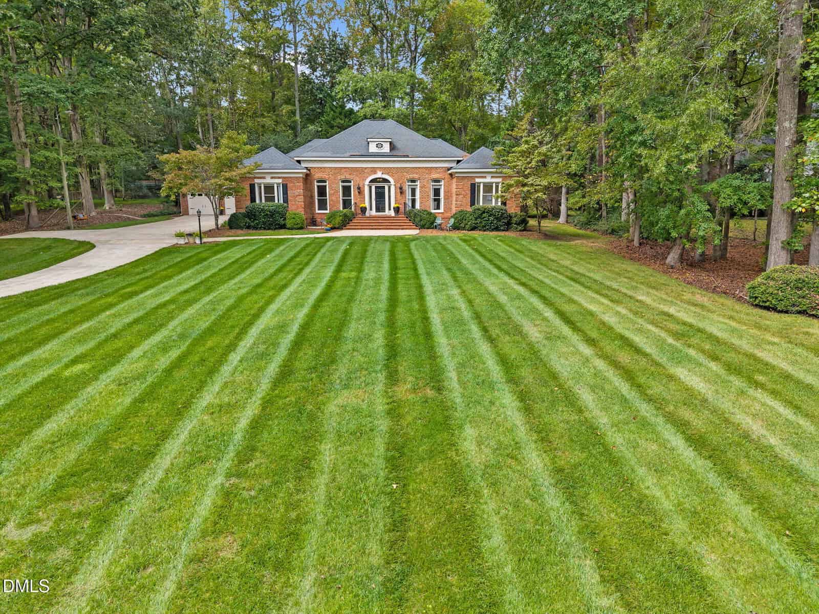 A large brick house with white trim sits at the end of a long, striped, freshly mowed lawn, surrounded by tall trees on both sides and a curved driveway on the left.