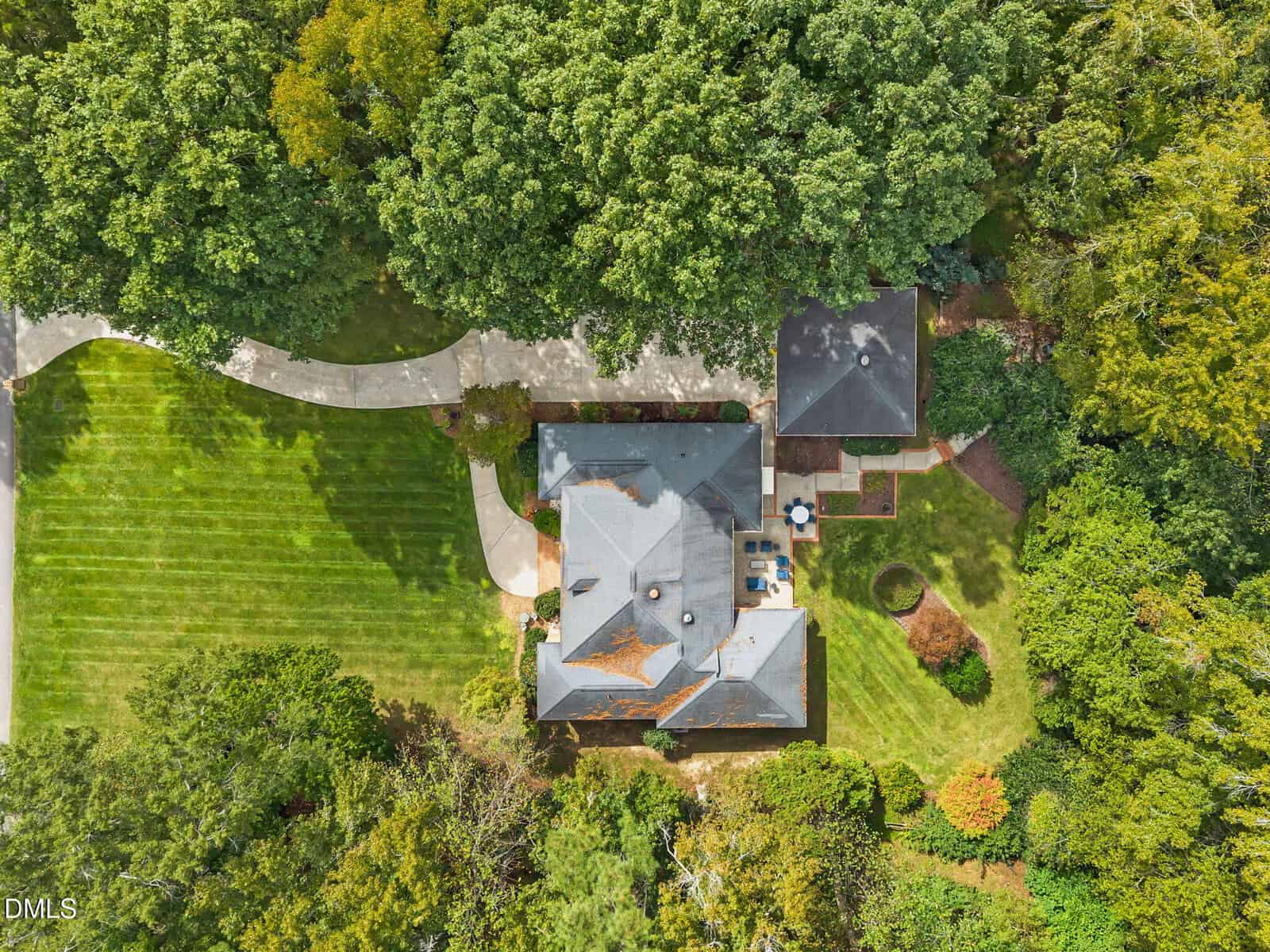 Aerial view of a large house with a gray roof, surrounded by dense trees, a manicured lawn, curved driveway, patio area, and various landscaped sections. The property appears well-maintained and bordered by greenery.