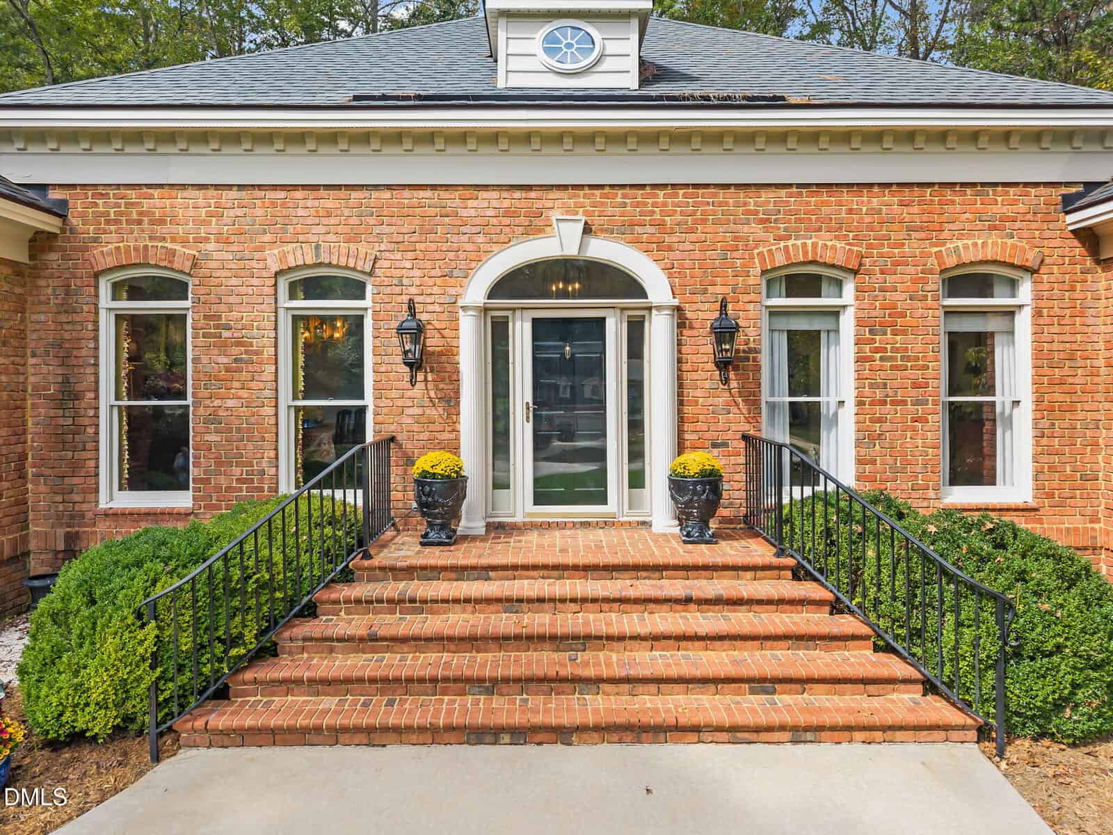 A brick house exterior with a central glass door framed by an arch, two black lantern-style lights, potted yellow flowers on each side of the entrance, and steps with black metal railings leading to the front door.