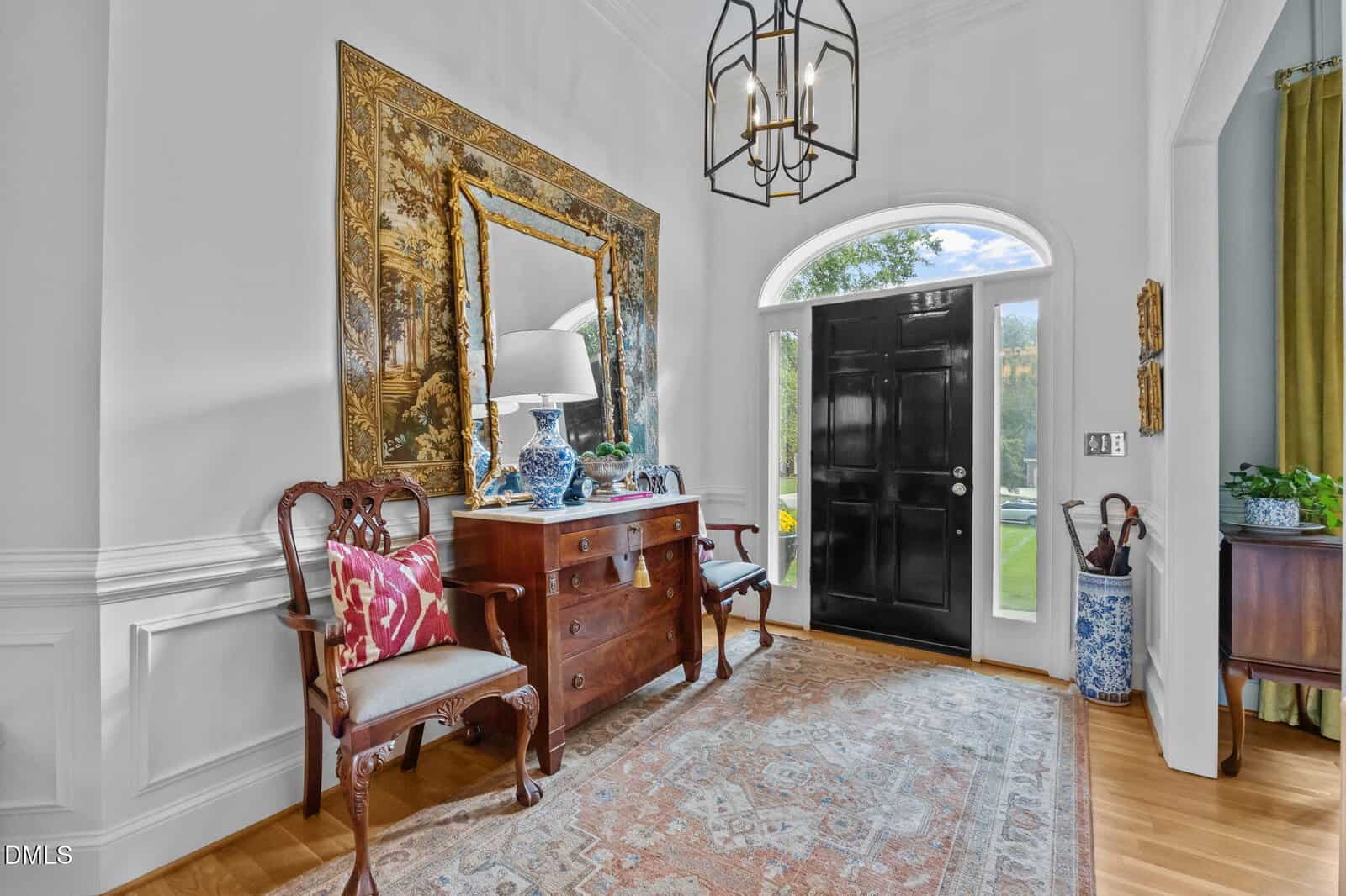 A bright entryway with a black front door, glass sidelights, and an arched window. A wooden console table, decorative mirror, chairs, and ornate rug furnish the space. A chandelier hangs from the ceiling.