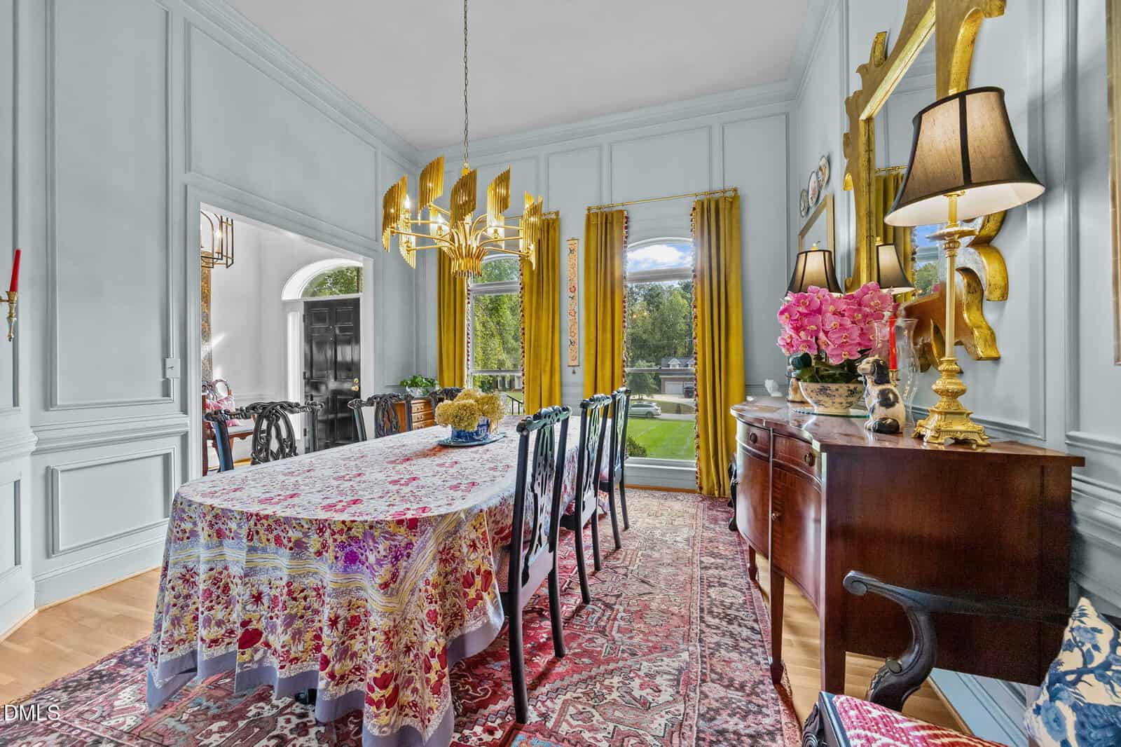 A formal dining room with a long table covered by a patterned cloth, dark chairs, a gold chandelier, a sideboard with lamps and flowers, yellow curtains, and a large window overlooking a garden.