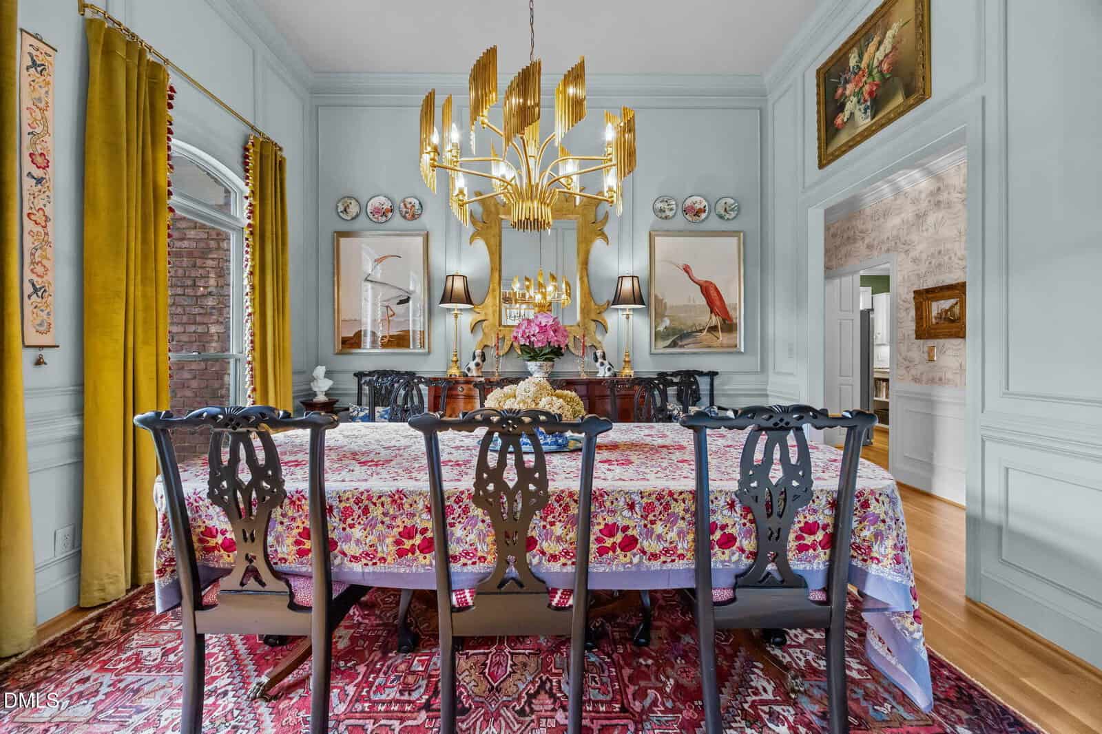 A dining room features a long table with a floral tablecloth, six dark wooden chairs, a gold chandelier, and a sideboard with lamps and flowers. Artwork and decorative plates hang on pale blue paneled walls.