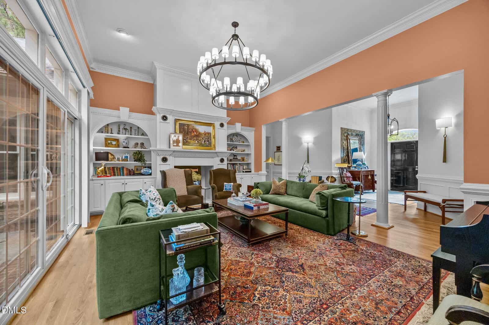 Spacious living room with green sofas, a colorful rug, built-in shelves, a grand chandelier, artwork above the fireplace, and large windows letting in natural light. Warm orange accents on the walls.
