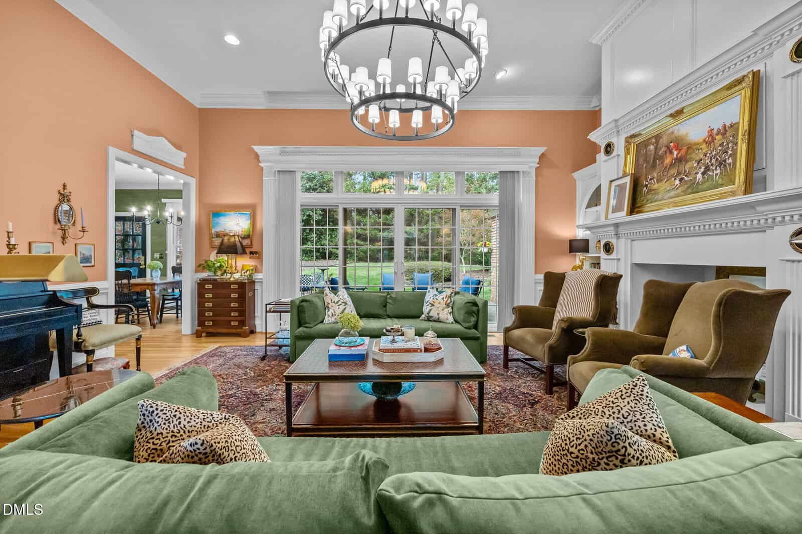 Elegant living room with green sofas, brown armchairs, a piano, large windows overlooking a garden, a chandelier, and a white mantle with artwork above the fireplace. Warm peach walls and classic decor create a cozy atmosphere.