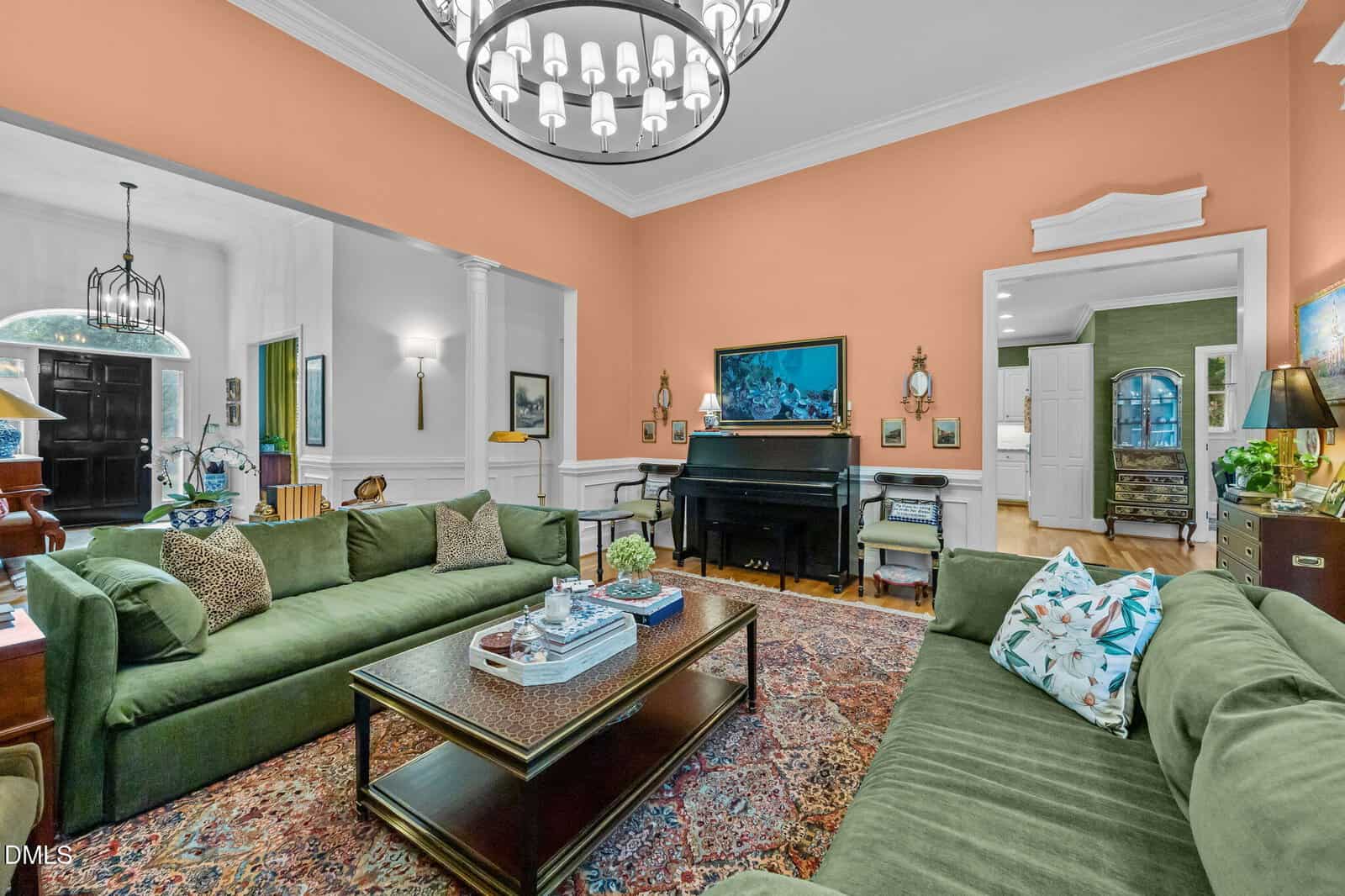 Elegant living room with two green sofas, a patterned rug, a black piano, peach-colored accent wall, chandelier, art, plants, books, and decorative pillows. Adjacent rooms visible, featuring white and green walls.