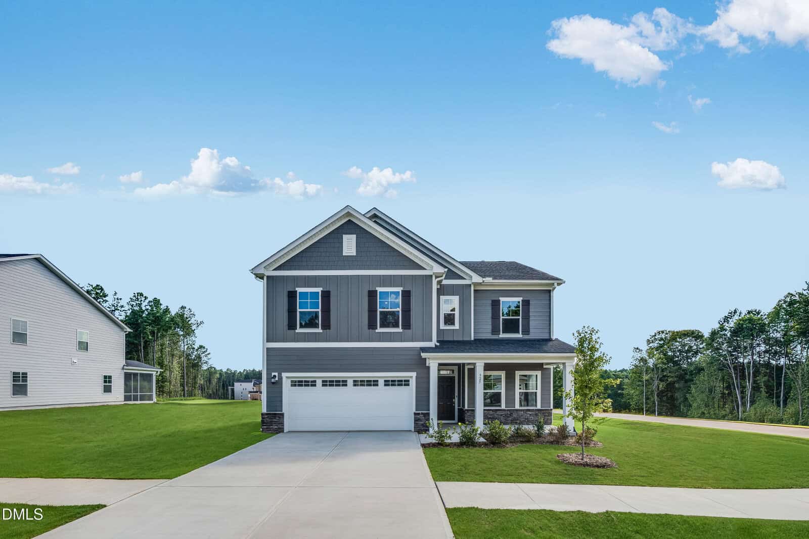 Two-story gray house with white trim and attached garage, set on a large green lawn with a driveway. The sky is blue with scattered clouds. Other homes and trees are visible in the background.