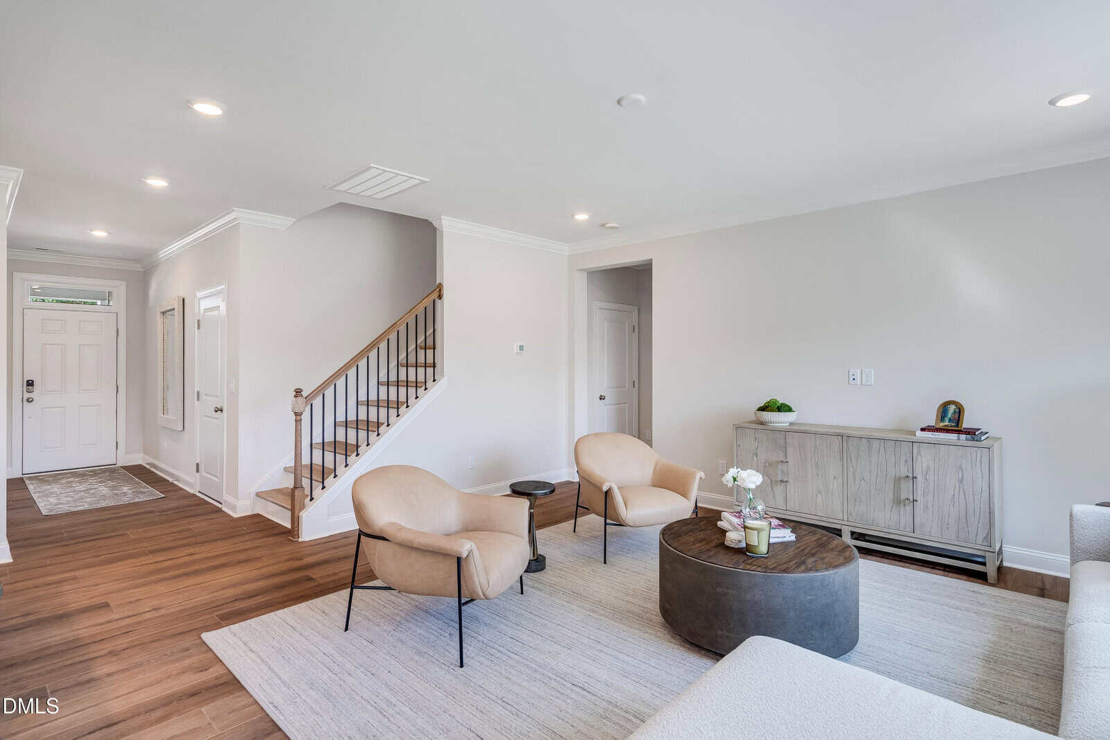 A bright living room with light walls, wood flooring, two tan chairs, a round coffee table, a sofa, a cabinet with decor, and a staircase leading upstairs. A door and hallway are visible in the background.