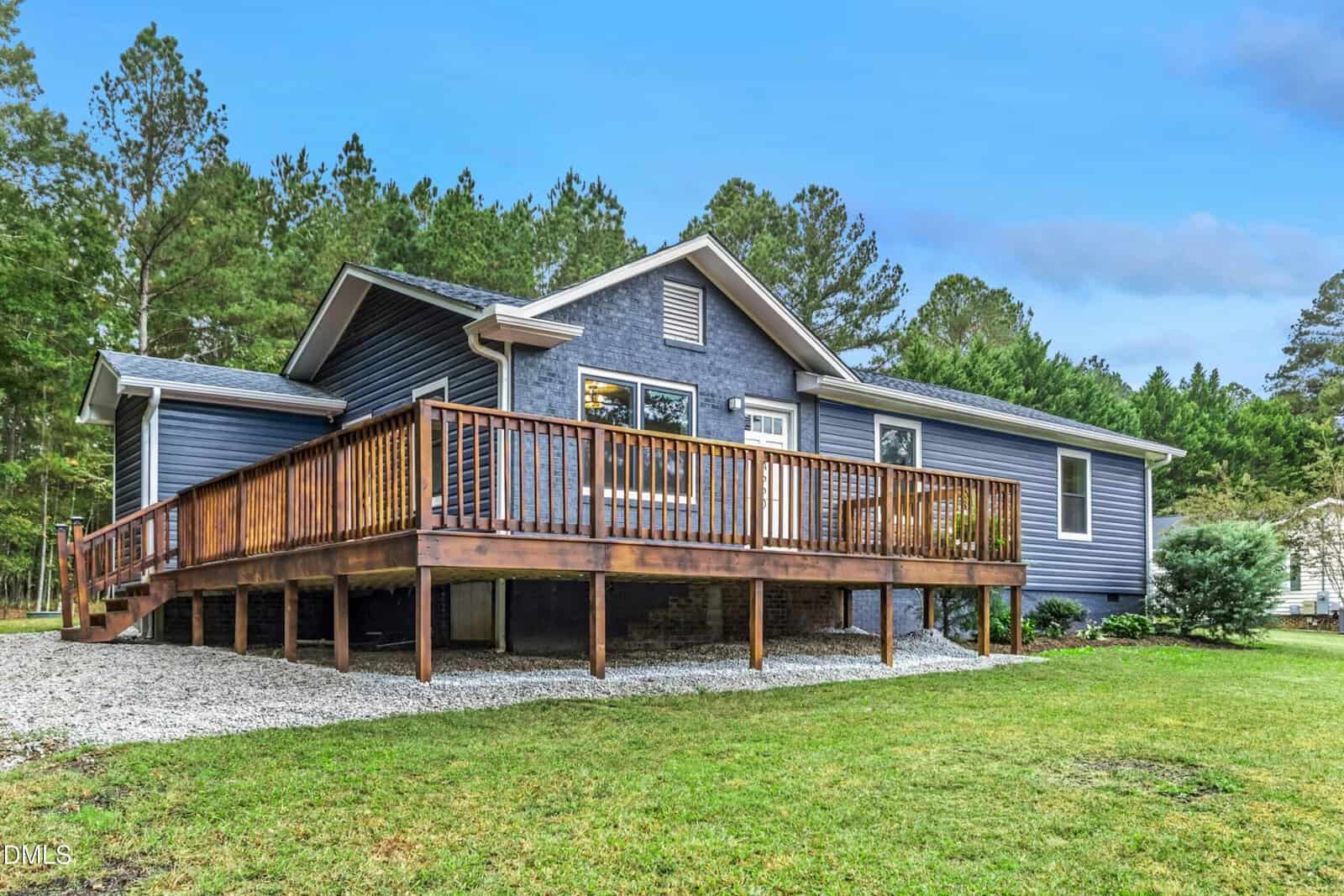 A single-story house with blue-gray siding and a large wooden deck, surrounded by grass, trees, and gravel, under a partly cloudy sky.