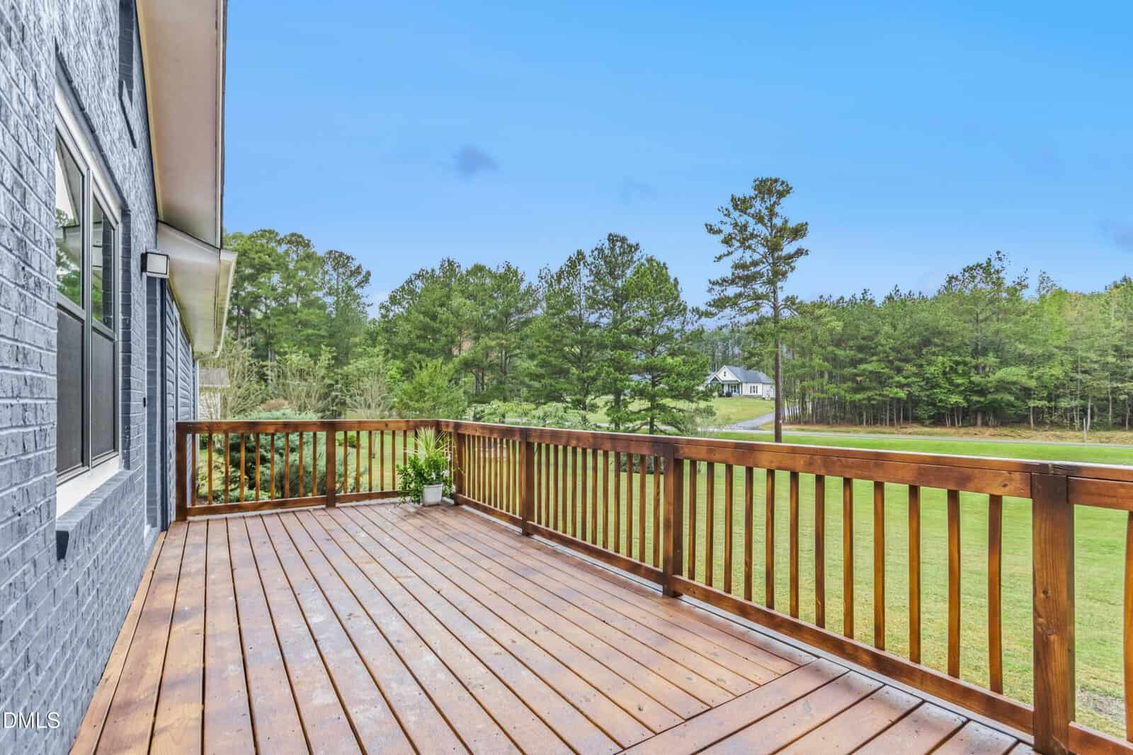 A wooden deck with a railing attached to a brick house overlooks a large grassy yard bordered by trees. A potted plant sits in one corner, and a house is visible in the distance. The sky is clear and blue.