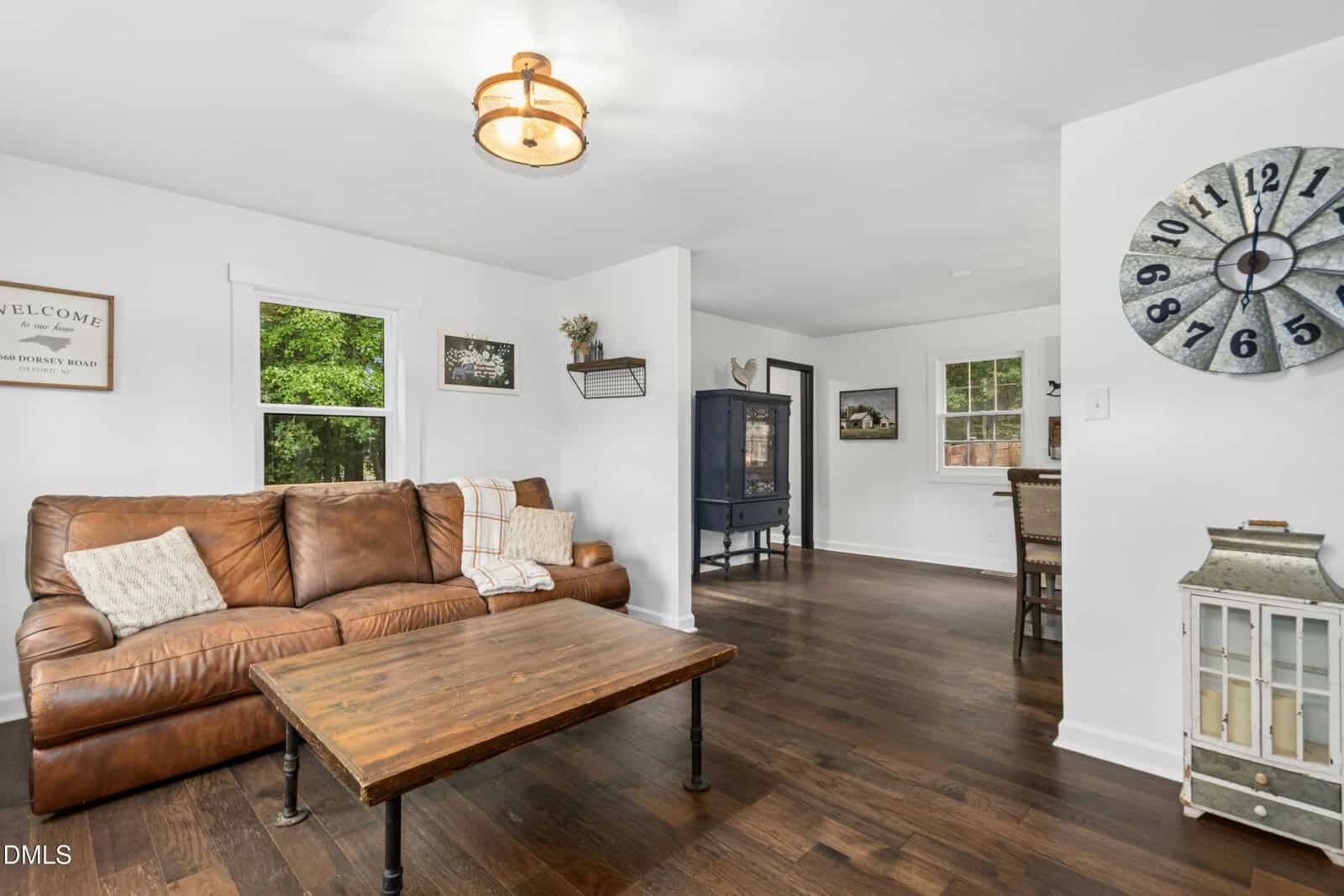 A living room with a brown leather sofa, wooden coffee table, white walls, dark wood floors, and a large wall clock. A window shows greenery outside, and a dining area is visible in the background.