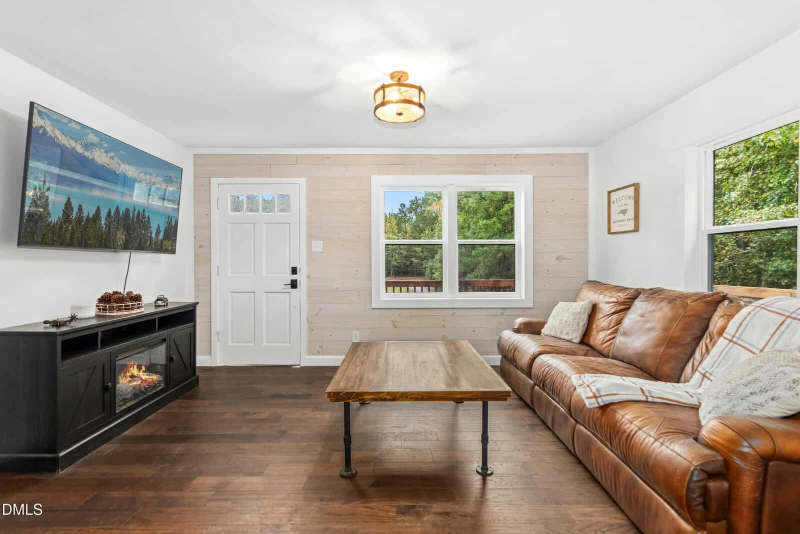 A living room with hardwood floors, a brown leather sofa, a wooden coffee table, a black fireplace TV stand, a wall-mounted TV, white walls, and two windows letting in natural light.