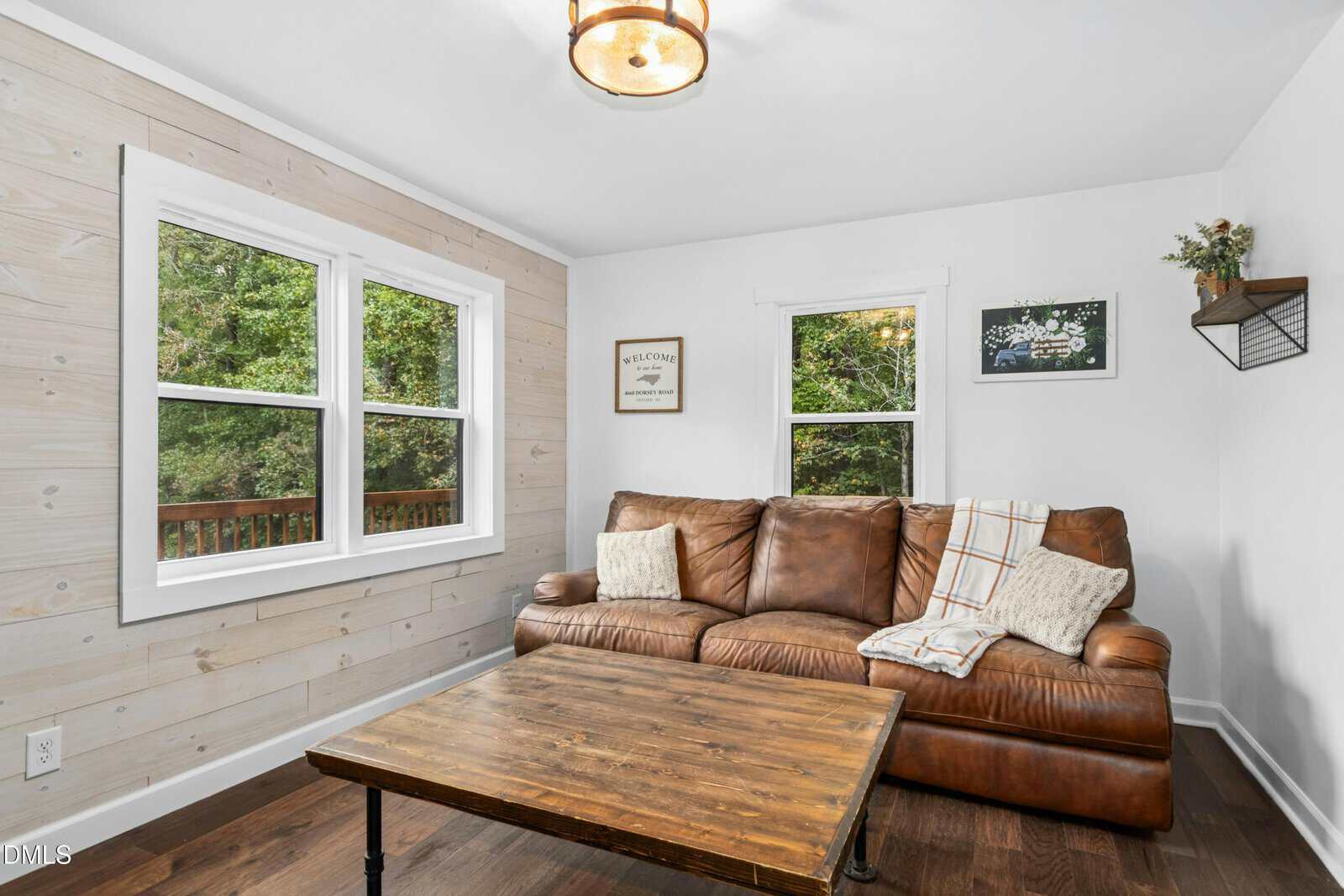 A small living room with a brown leather couch, a wooden coffee table, white walls, and two windows showing green trees outside. Throw pillows and a blanket are on the couch, and two pictures hang on the wall.