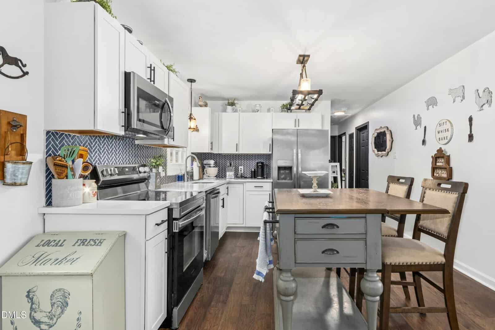 Modern kitchen with white cabinets, stainless steel appliances, blue tile backsplash, and a wooden dining table with four chairs. Decorative accents and kitchen items are displayed on counters and walls.