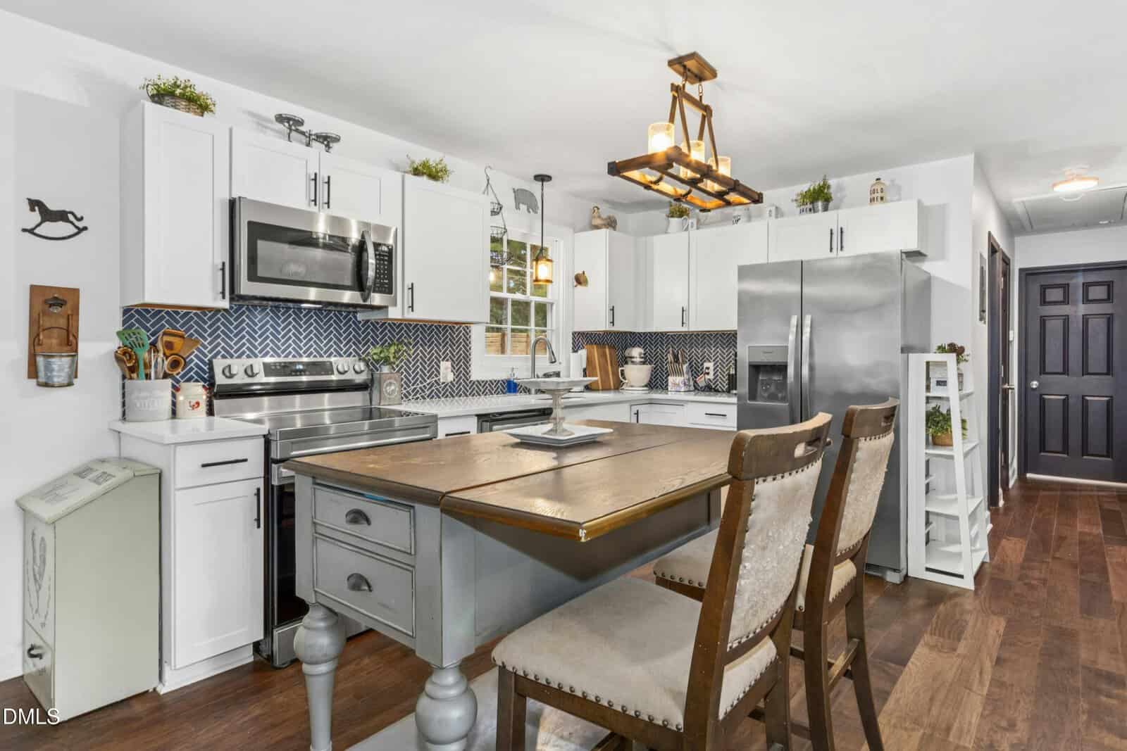 A modern kitchen features white cabinets, stainless steel appliances, a central island with two chairs, a patterned blue backsplash, wooden floors, and decorative plants on top of the cabinets.