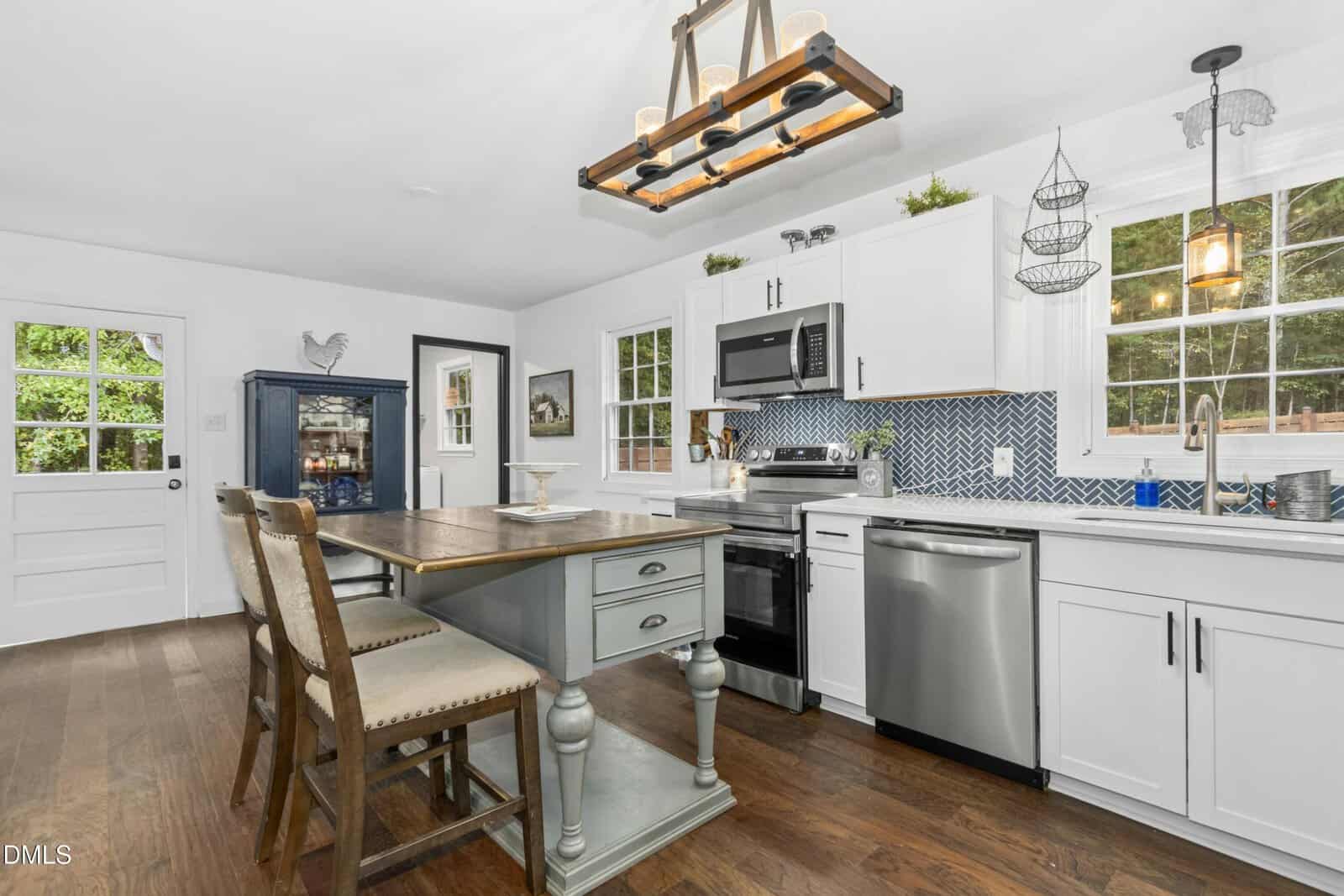 Bright kitchen with white cabinets, stainless steel appliances, a patterned blue backsplash, large windows, a wooden dining table with two chairs, light fixture above, and hardwood floors. Door and hutch visible in the background.