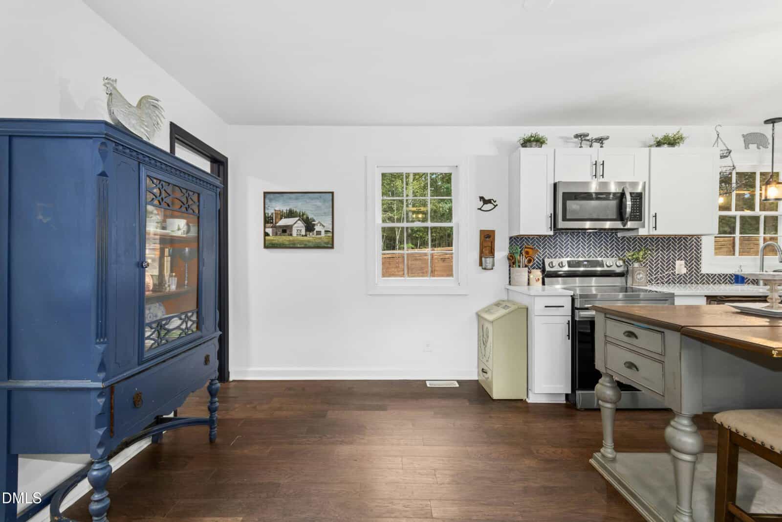 A kitchen with white cabinets, stainless steel appliances, blue and white backsplash, a dark blue hutch on the left, a central island, hardwood floors, and a window above a small beige cabinet.