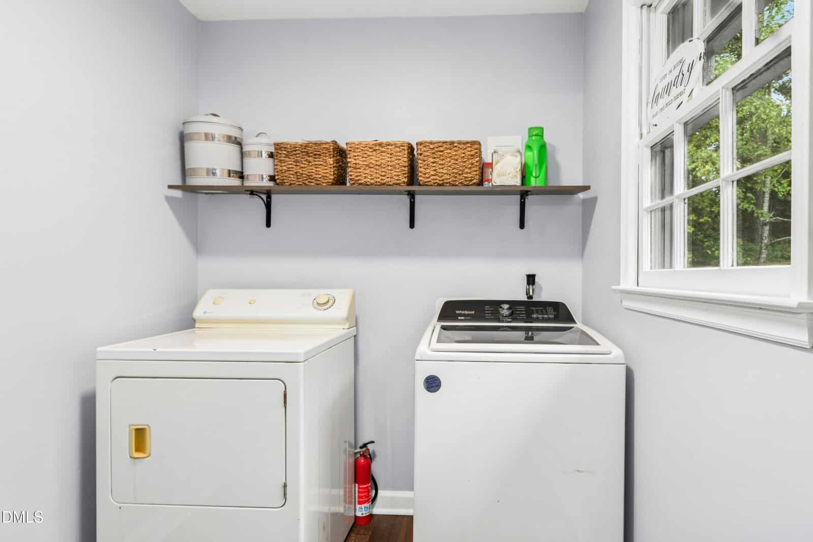 A laundry room with a dryer and a washing machine, a shelf above holding baskets and containers, a fire extinguisher on the floor, and a window on the right letting in natural light.
