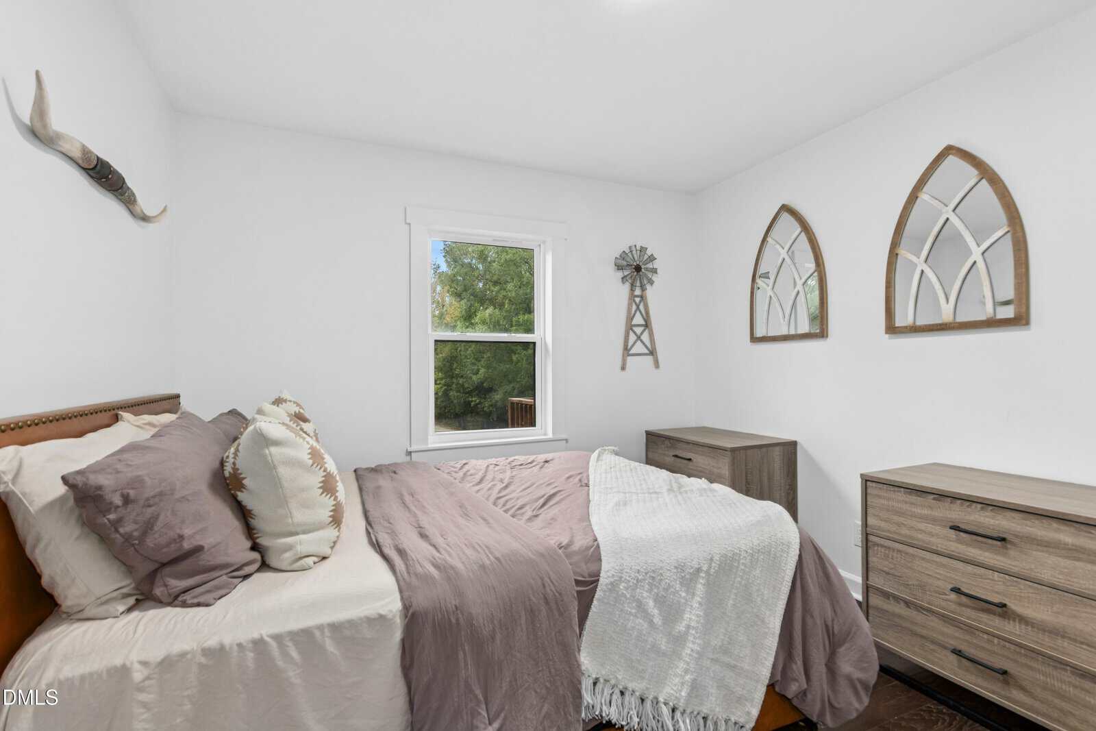 A cozy bedroom with a bed in soft beige and mauve bedding, two dressers, wall decor including mirrors and a windmill piece, and a window showing green trees outside.