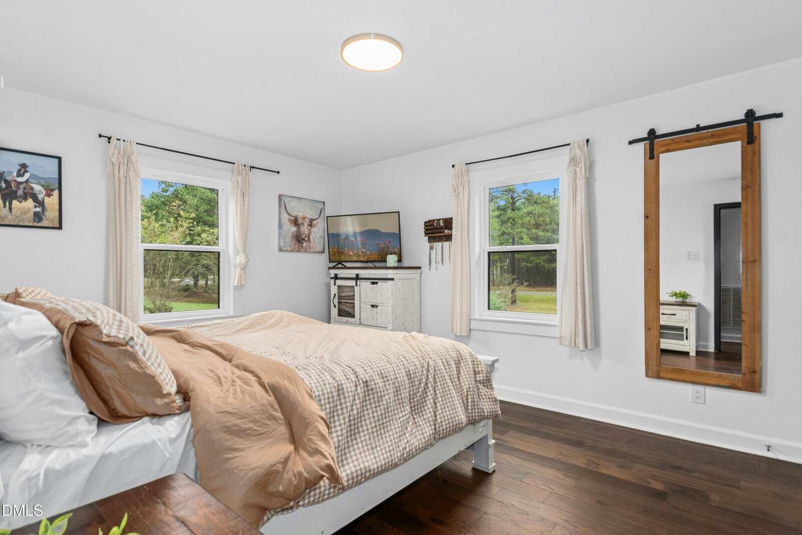 Bright bedroom with a large bed, white walls, wood floors, three windows with curtains, a wall mirror, dresser, and animal-themed artwork. Green trees are visible through the windows.