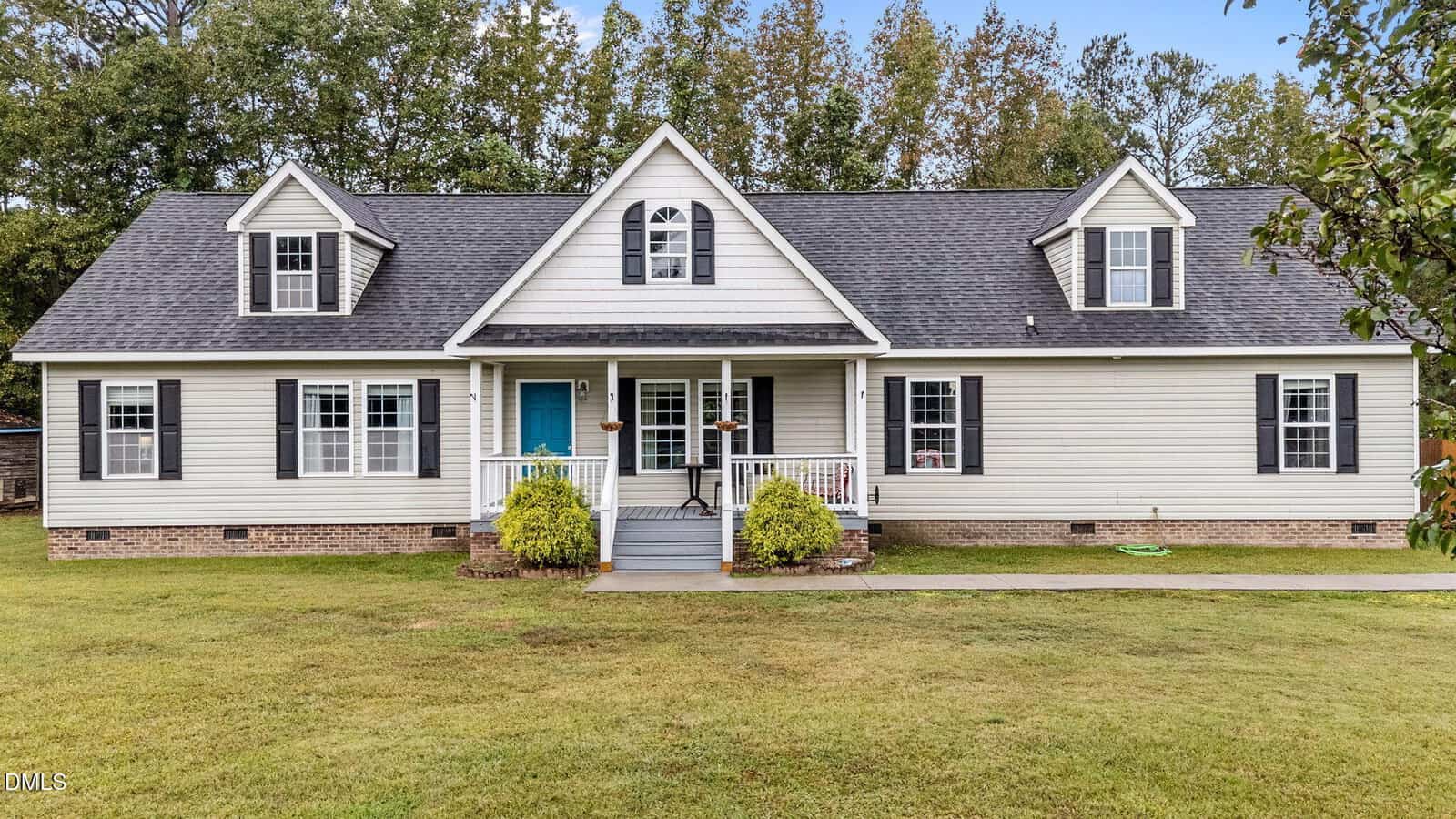 Single-story house with white siding, dark shutters, two dormer windows, a front porch with railings, a blue front door, and small bushes by the entrance. The lawn is neatly mowed and trees are in the background.