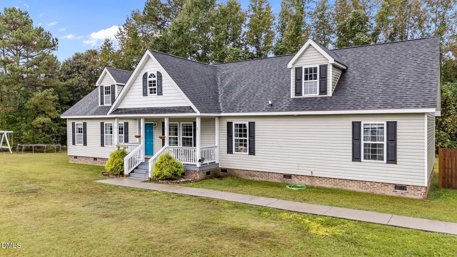 A beige, single-story house with a dark roof, white trim, and black shutters sits at 2253 Pete Smith Road in Louisburg. It features two dormer windows, a front porch with columns, and a blue door. The yard is grassy with trees and a sidewalk leading to the entrance.