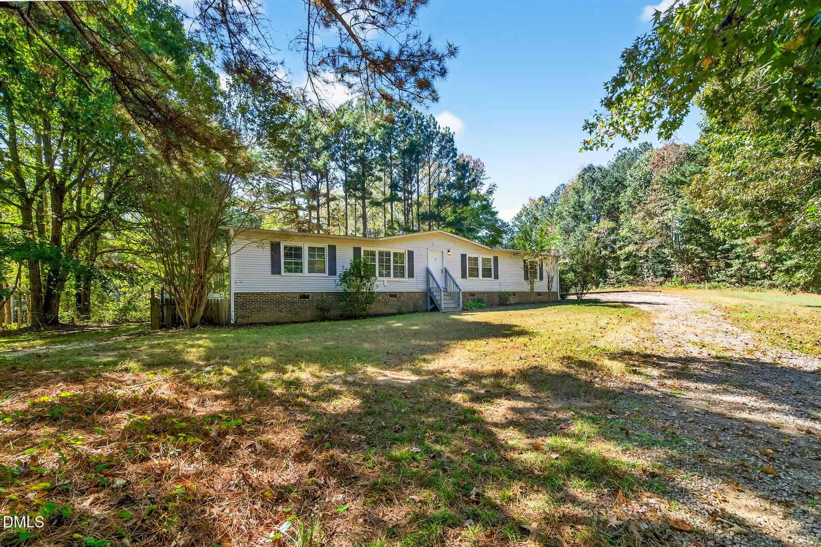 Single-story white house with blue steps and shutters, surrounded by trees and a grassy yard at 517 Mitchiner Road, Franklinton. A gravel driveway curves from the front right toward the house. The scene is lit by bright sunlight.