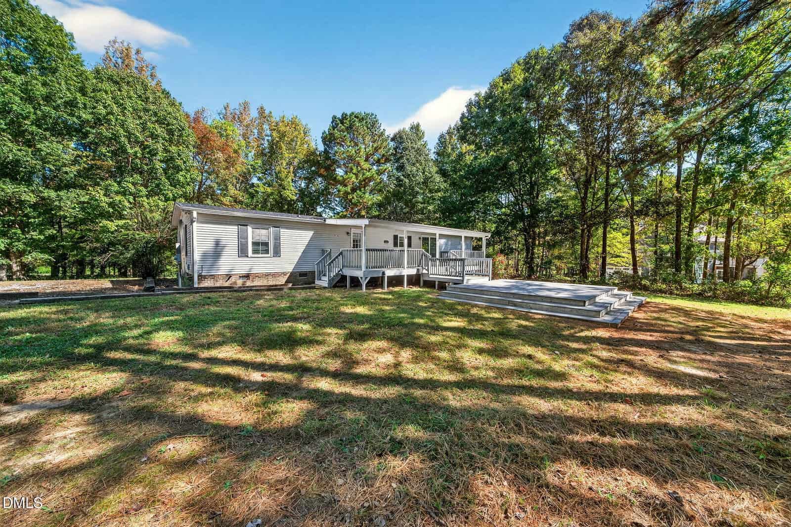 A single-story white manufactured home with a covered front porch and large wooden deck, surrounded by trees and grass under a blue sky with scattered clouds.