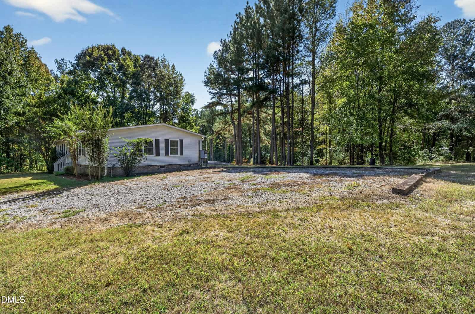 A single-story house with light siding sits next to a gravel driveway, surrounded by tall trees and grass under a blue sky with scattered clouds.