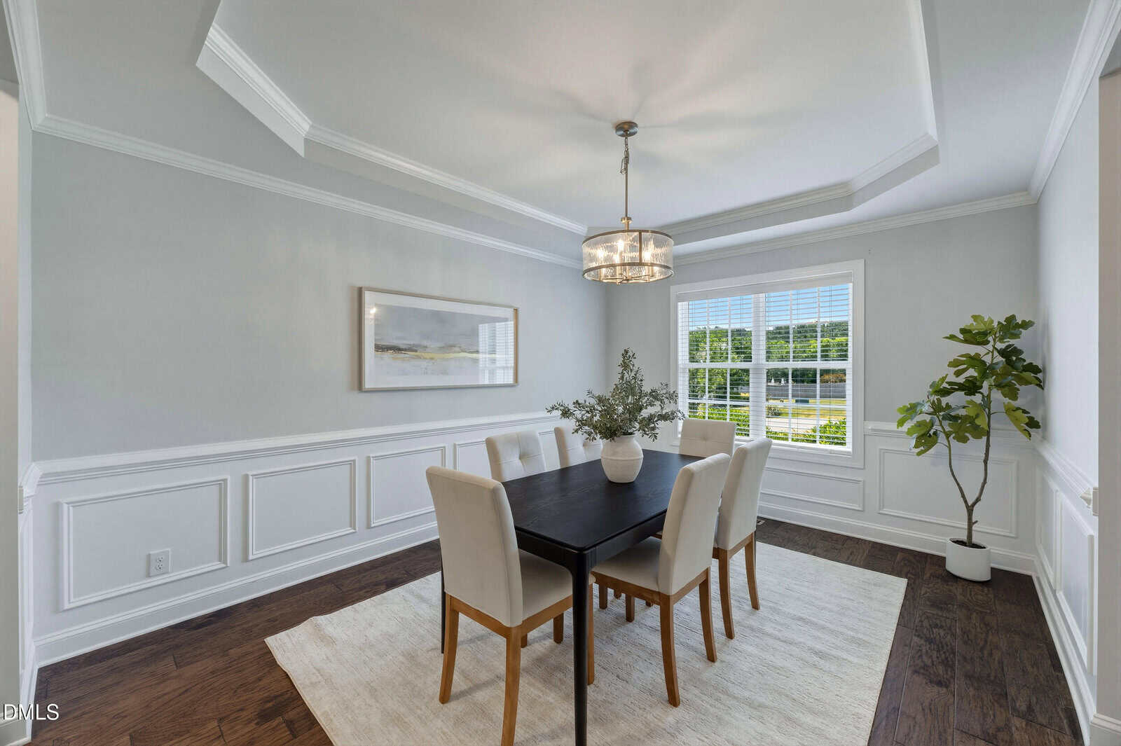 A bright dining room with a black table, six beige chairs, a chandelier, a large window, a potted plant, a framed picture on the wall, white paneled walls, and dark wood floors with a light rug.