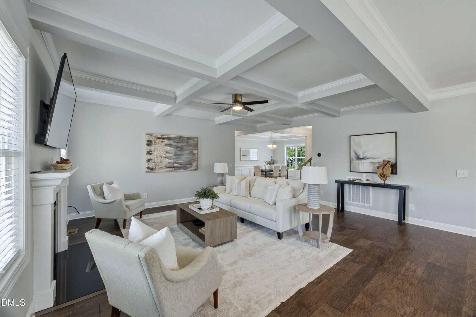 Bright living room with coffered ceiling, white sofa, two armchairs, coffee table, rug, wall art, and wood floors. Mounted TV above fireplace and a console table with decor against the back wall.