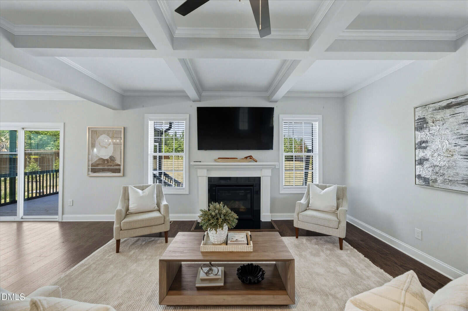A bright living room with two armchairs, a coffee table, and a rug facing a fireplace with a mounted TV above it. Large windows let in natural light, and artwork decorates the light-colored walls.