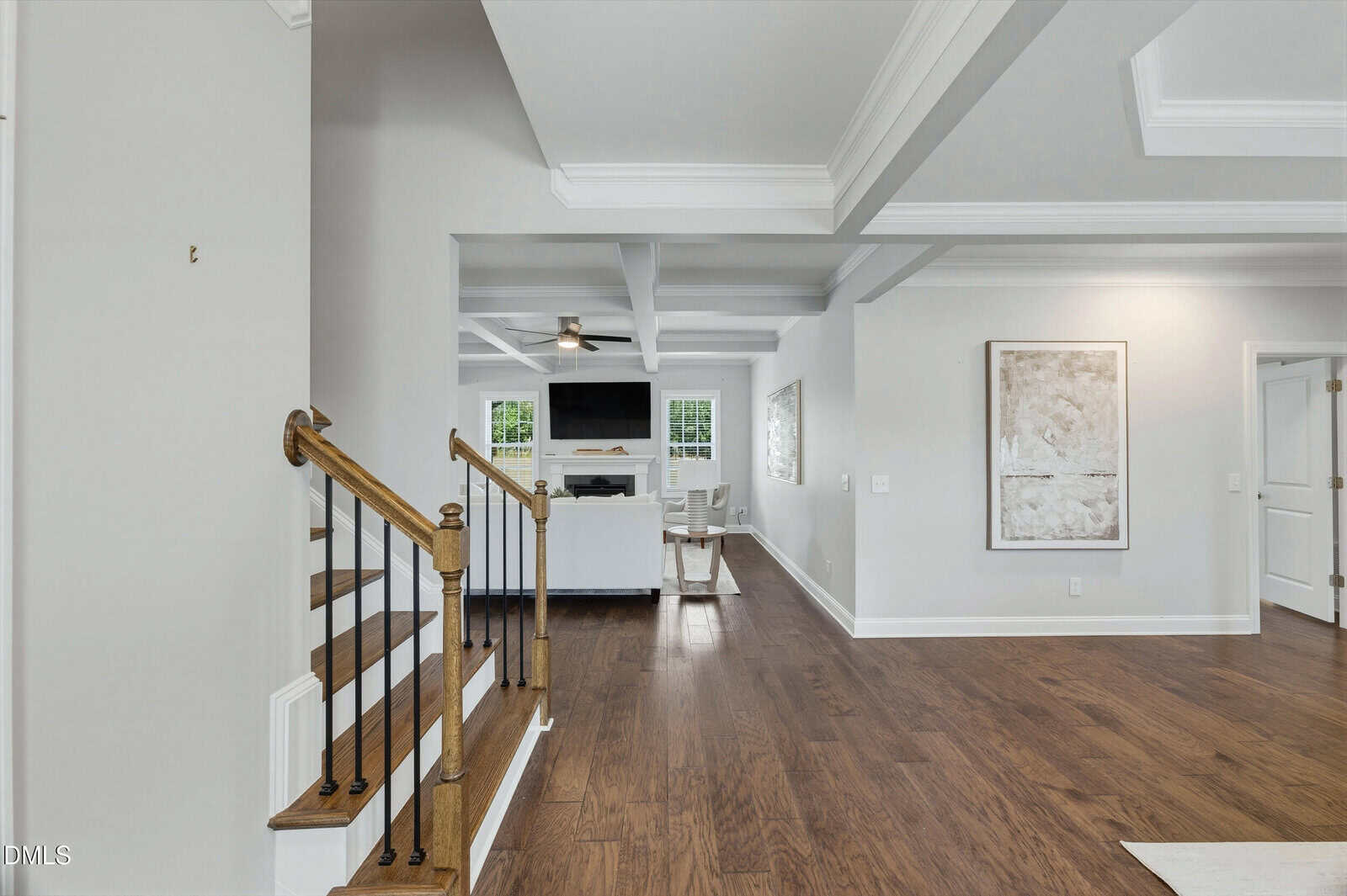 Spacious, modern living area with hardwood floors, neutral walls, a staircase with wooden handrail and black balusters, white coffered ceiling, and a sitting area with a TV and windows in the background.