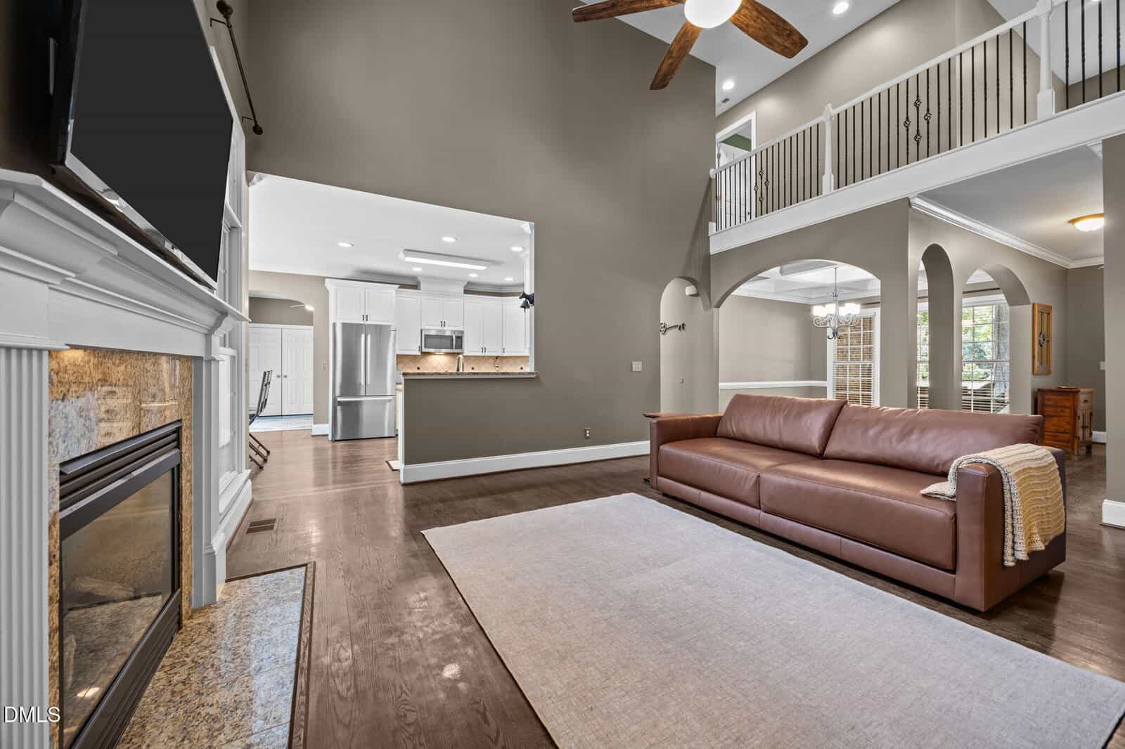 Spacious living room with high ceilings, brown leather sofa, large area rug, fireplace, ceiling fan, and open view to the kitchen and dining areas with white cabinetry and stainless steel appliances. Balcony overlooks the space.