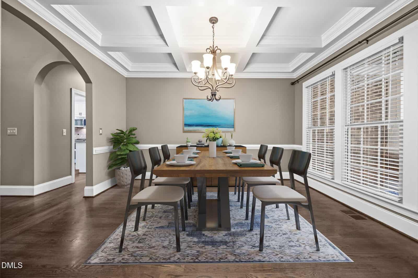 A spacious dining room with a long wooden table set for eight, modern chairs, a decorative rug, chandelier, large windows with blinds, beige walls, and a blue seascape painting above a side table with flowers.