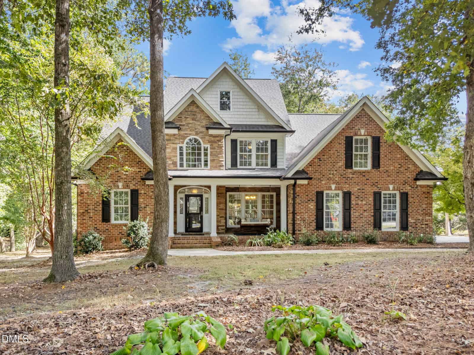 A two-story brick house with black shutters, multiple large windows, and a front porch at 5916 2 Pines Trail in Wake Forest, set among tall trees and surrounded by a dirt yard with some greenery in the foreground.