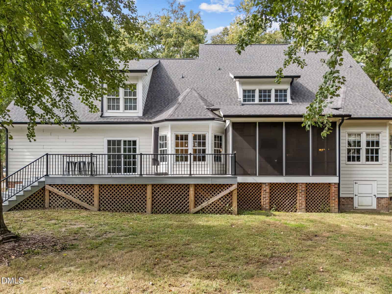 A two-story white house with a dark roof, featuring a raised wooden deck with railing, screened porch, and multiple windows, set in a grassy yard with trees in the background.