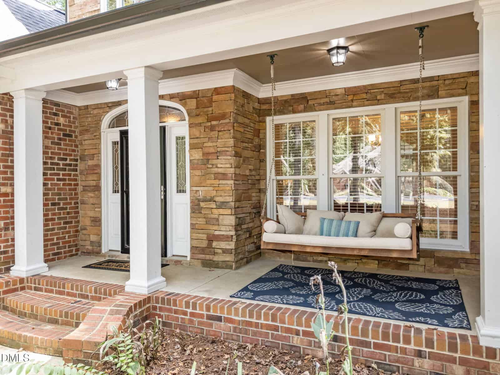 A brick and stone front porch with white columns, a glass double door entrance, a blue patterned rug, and a wooden porch swing with cushions and pillows.