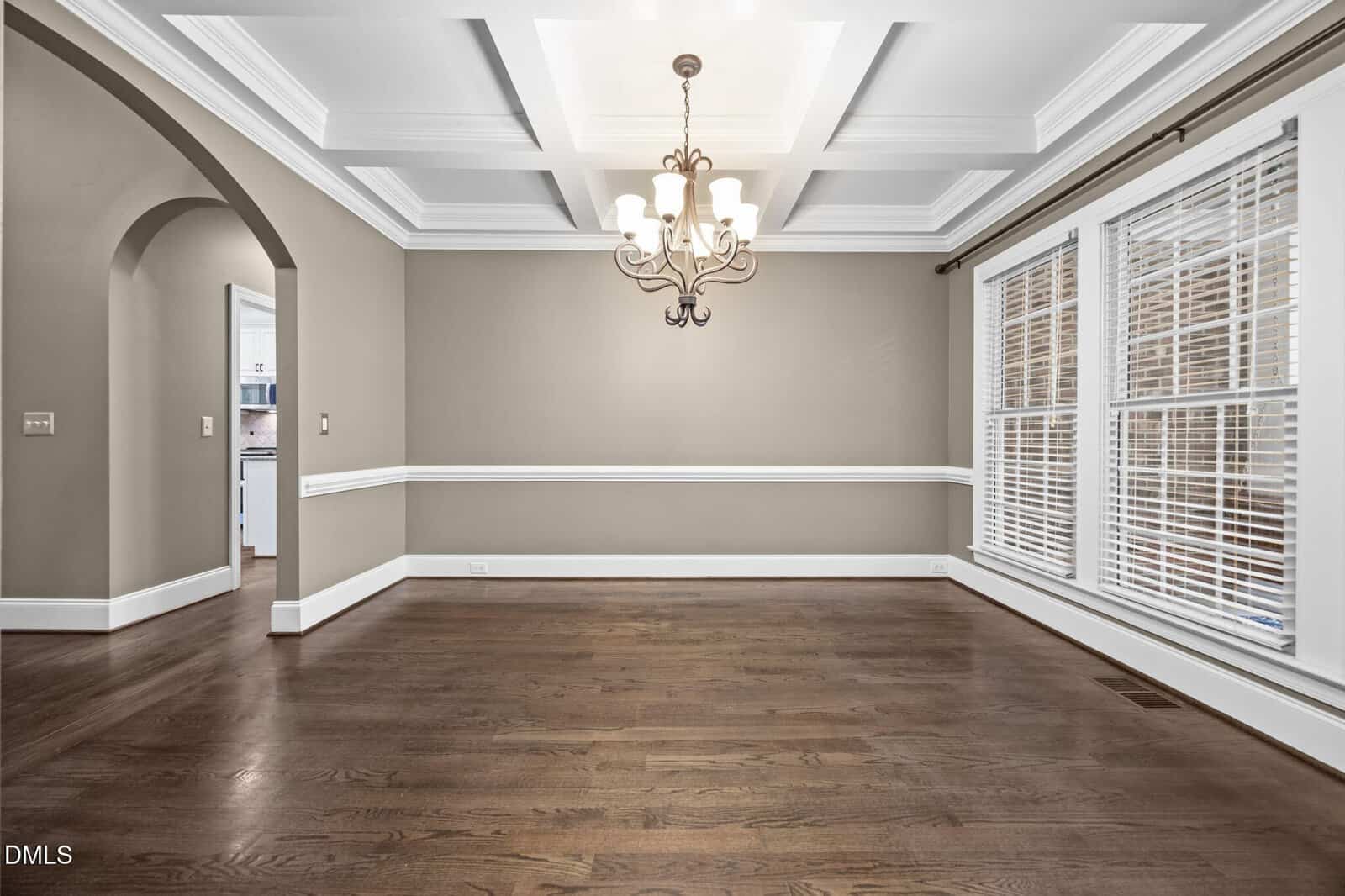 Spacious, empty dining room with hardwood floors, coffered ceiling, chandelier, crown molding, and large windows with white blinds. An arched doorway leads to another room.