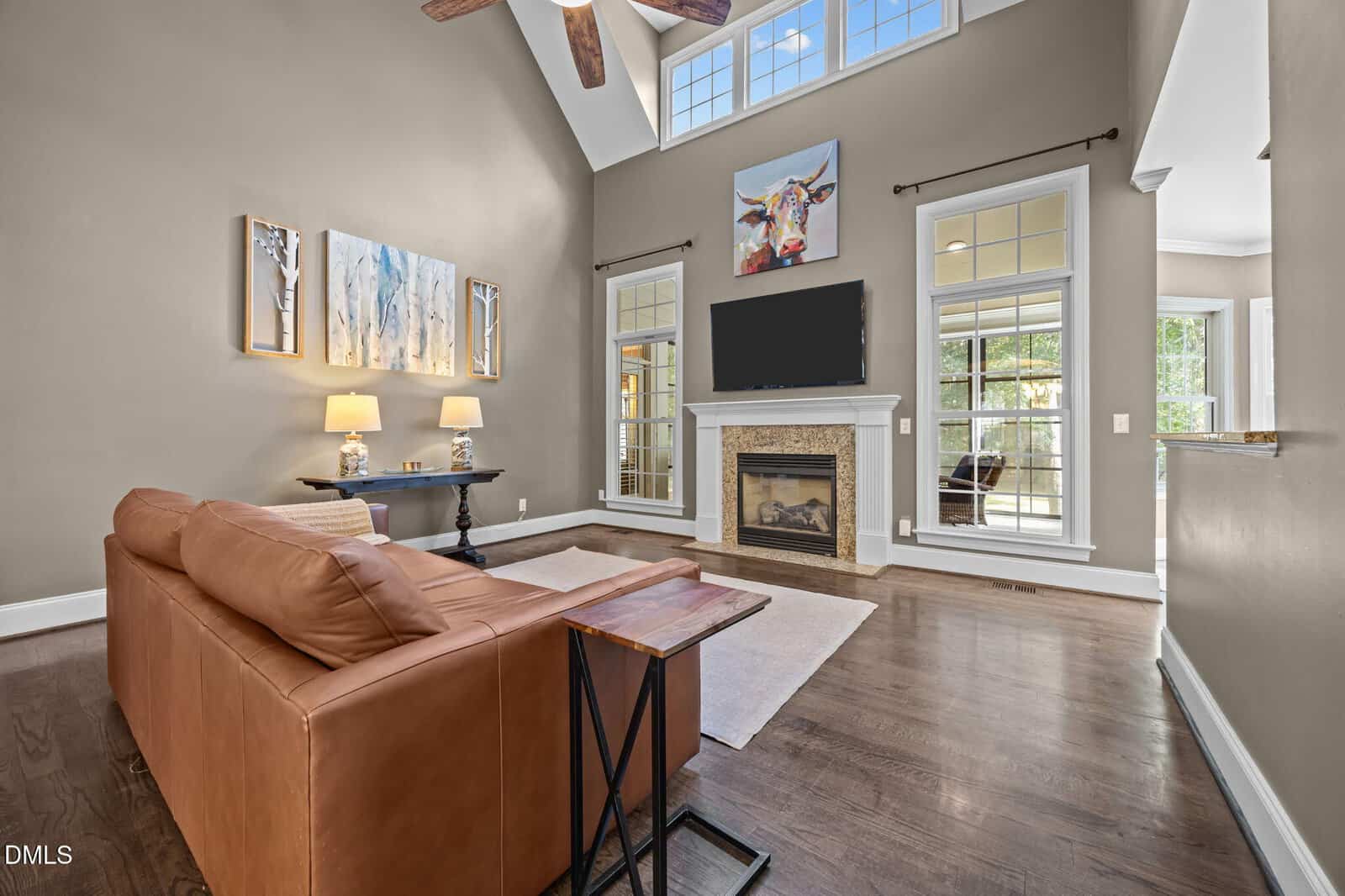 A modern living room with high ceilings, large windows, a brown leather sofa, wall art, a fireplace with a TV above it, and hardwood floors. Natural light brightens the spacious, open area.