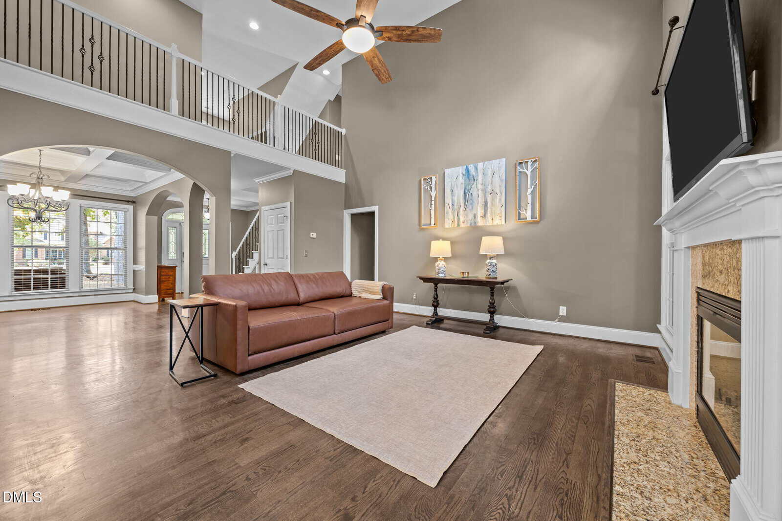 Spacious living room with high ceilings, brown hardwood floors, a brown leather sofa, a large area rug, a wall-mounted TV above a white fireplace, and a table with two lamps beneath abstract wall art.