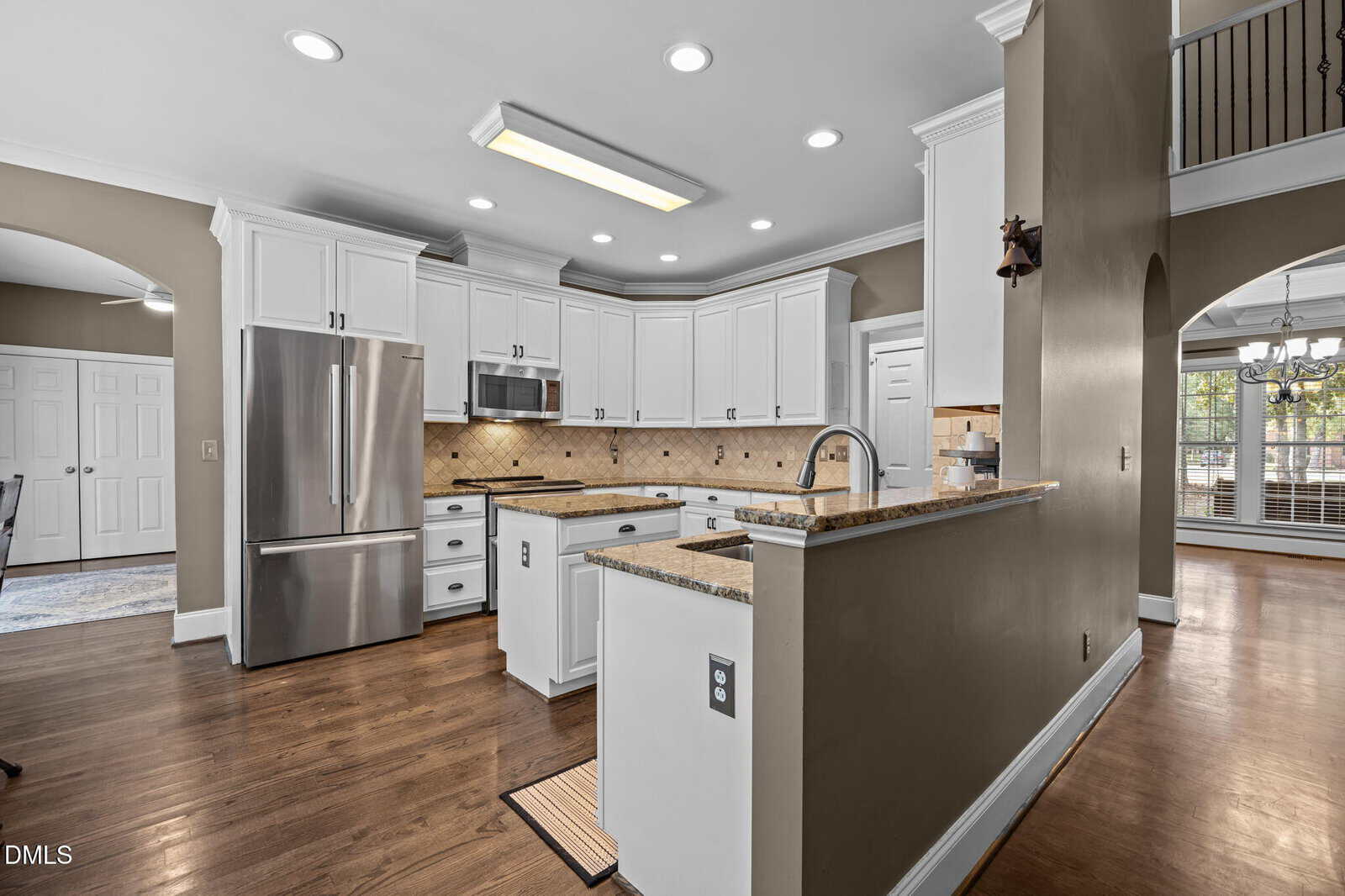 Modern kitchen with white cabinets, stainless steel appliances, granite countertops, wood floors, and recessed lighting. An arched doorway leads to a dining area with large windows.