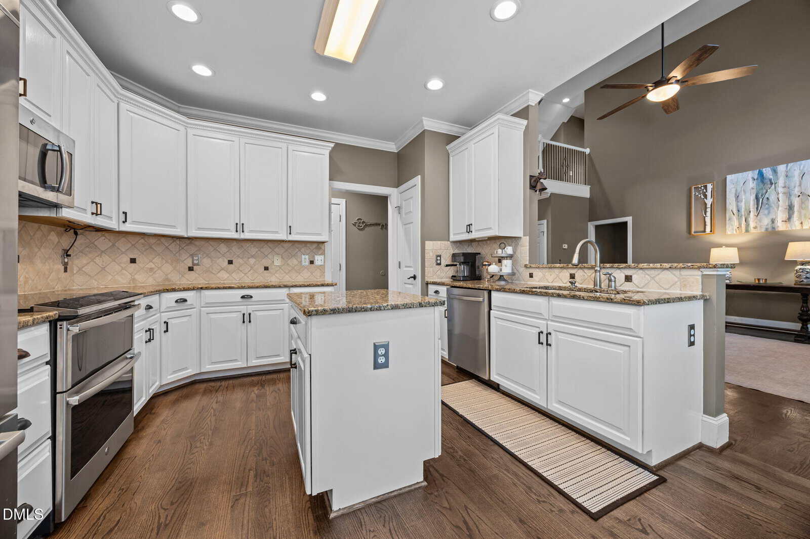 Spacious modern kitchen with white cabinets, stainless steel appliances, granite countertops, tile backsplash, and wood floors. An island and a sink are visible, with an open view to the living area and high ceilings.