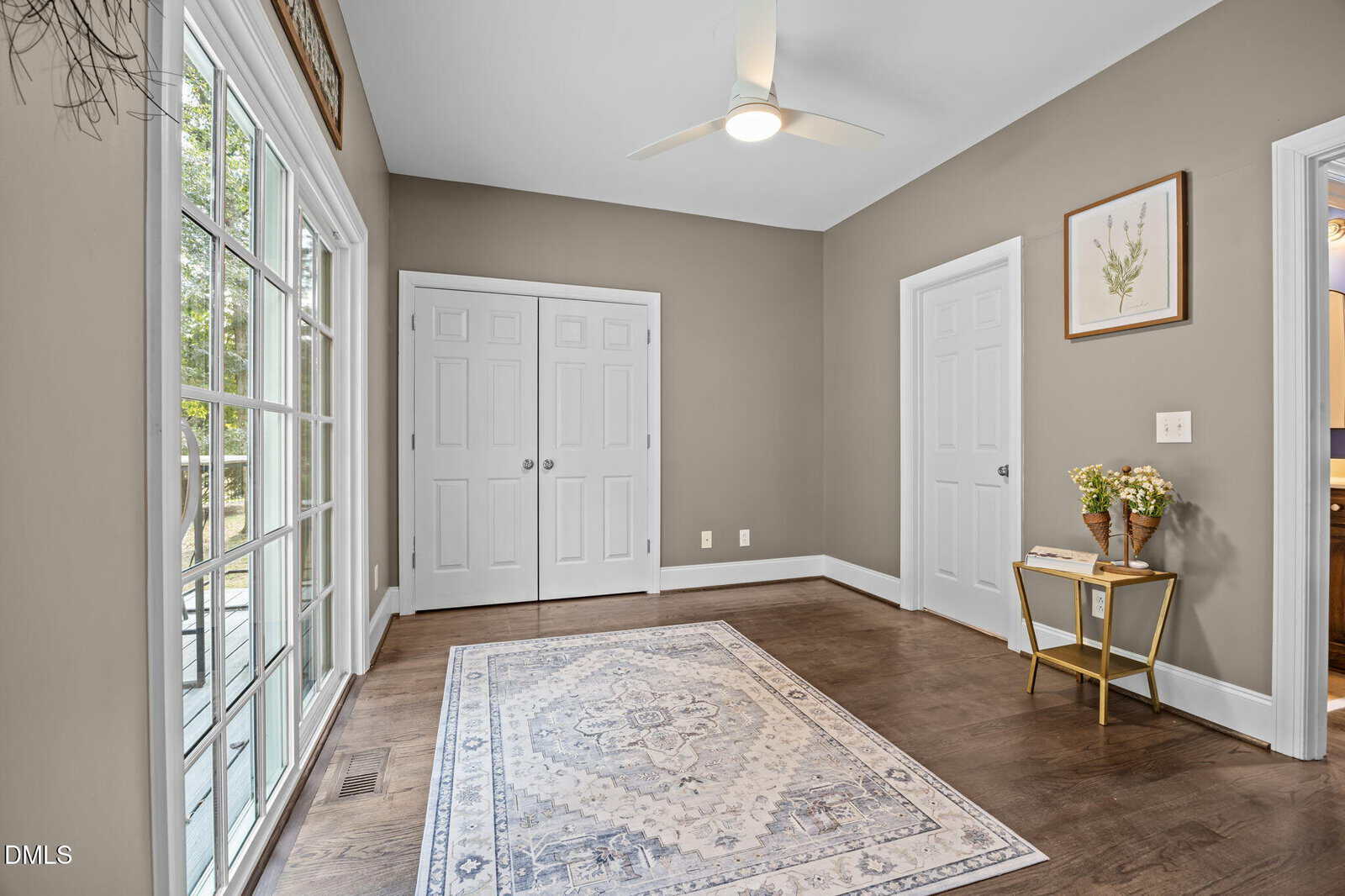 Bright room with large windows, a ceiling fan, double closet doors, and a patterned rug on dark wood floors. A small side table with flowers and framed art decorates the corner.
