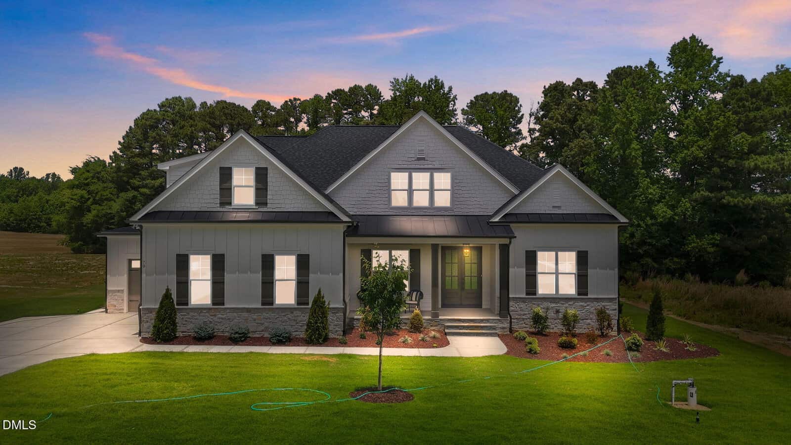 A modern, two-story house at 70 Firefly Lane, Youngsville features gray siding, stone accents, and a well-manicured lawn. Lit up at dusk, it sits amid small shrubs and trees with a stunning sunset sky in the background.