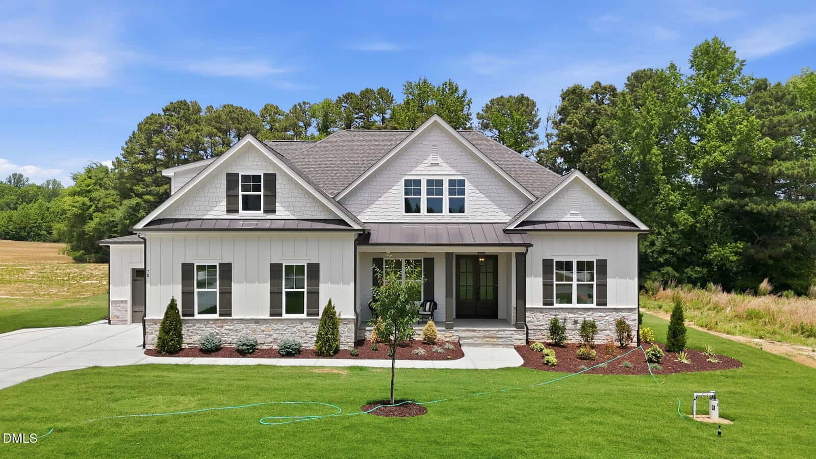 Modern two-story house with white siding, stone accents, and a gray roof, set on a large green lawn with landscaped shrubs and trees, surrounded by woods under a blue sky.