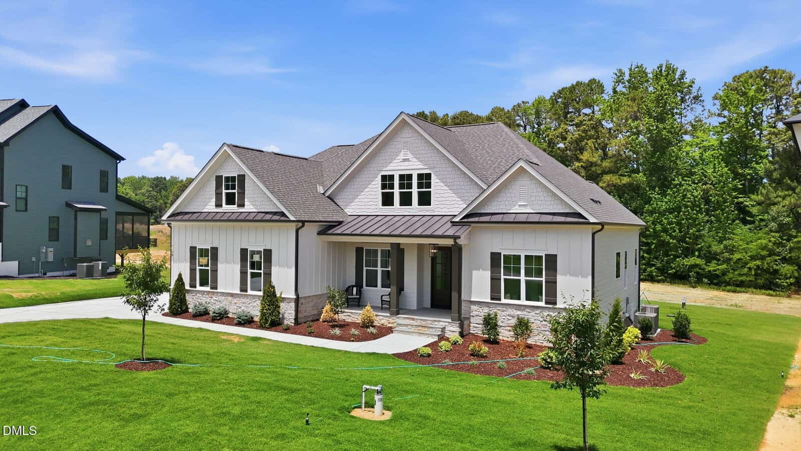 A modern two-story house with gray siding, white trim, a front porch, and manicured landscaping sits on a large, green lawn with neighboring houses and trees in the background under a blue sky.