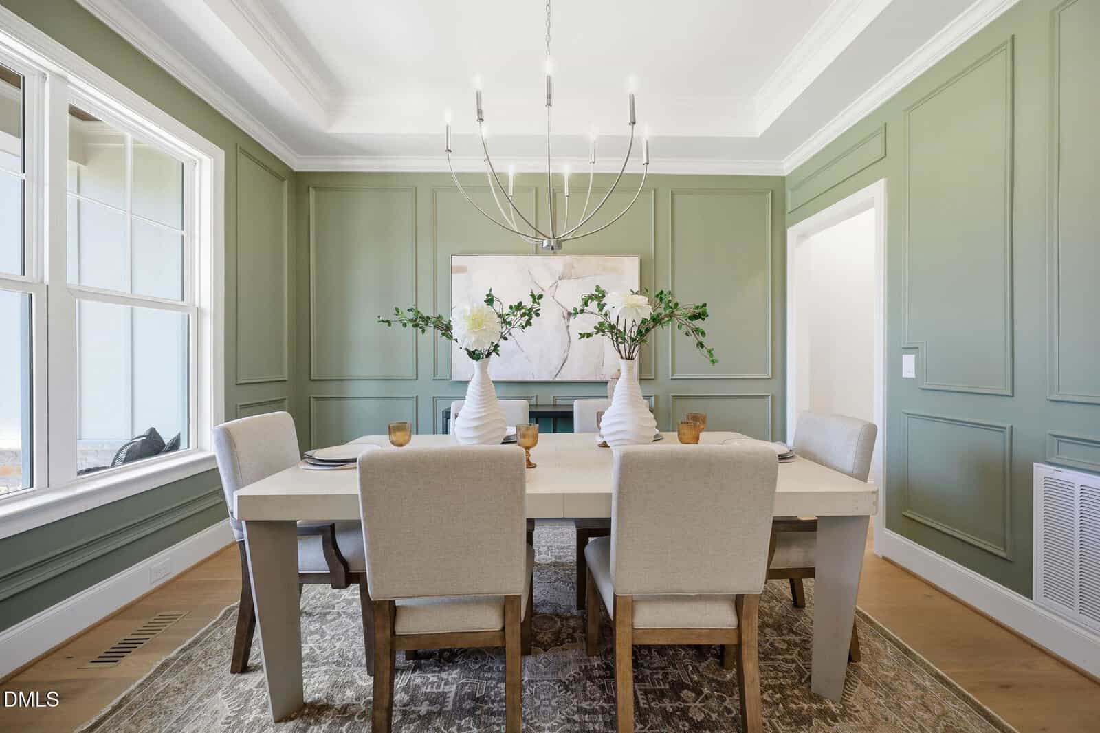 Elegant dining room with green paneled walls, a large white table, six beige upholstered chairs, modern chandelier, and decorative vases with greenery. Sunlight streams through tall windows on the left.