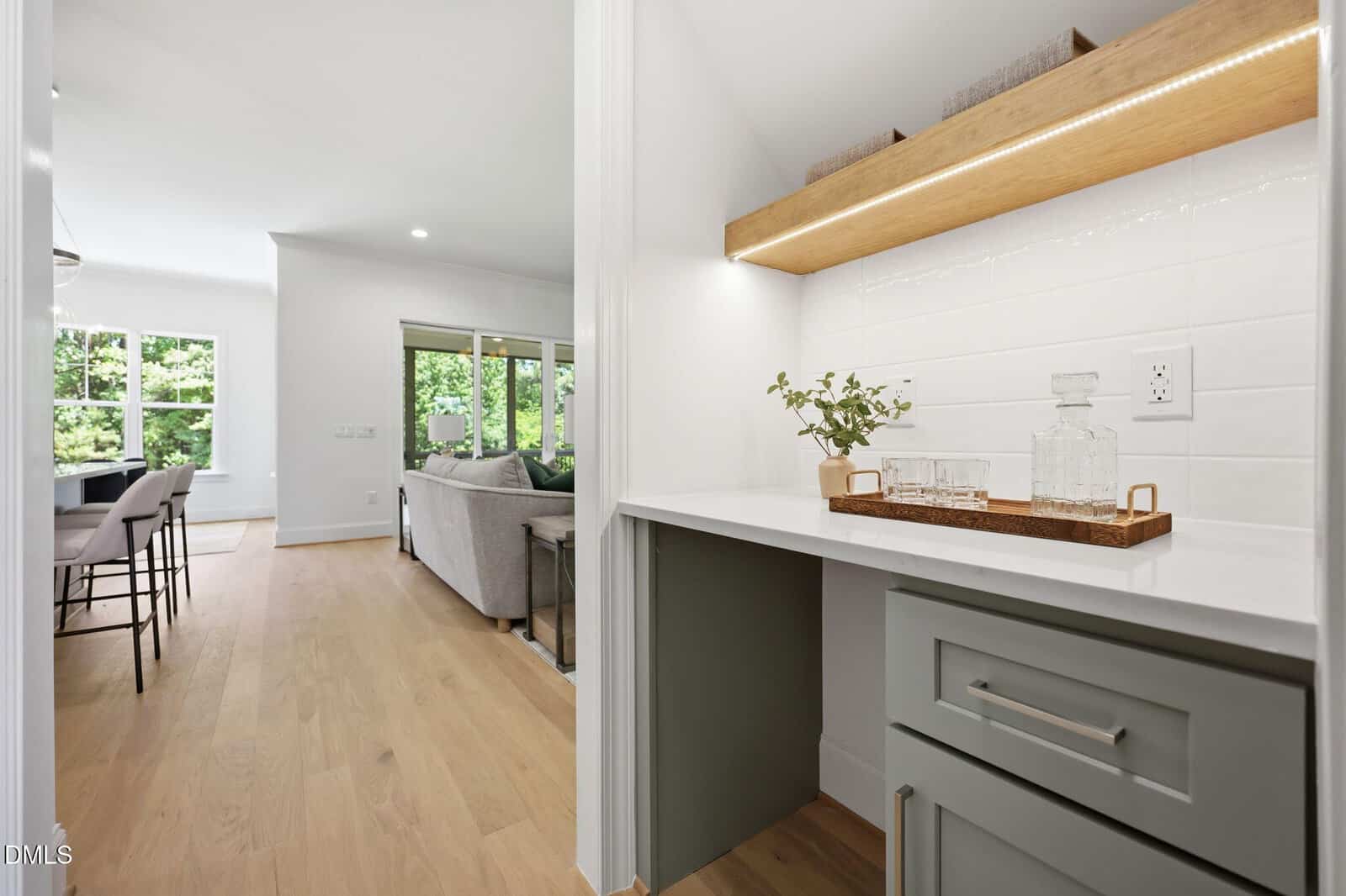 Modern home interior with a small bar area featuring glassware and a plant, light wood floors, neutral walls, and an open living and dining space with large windows and greenery outside.